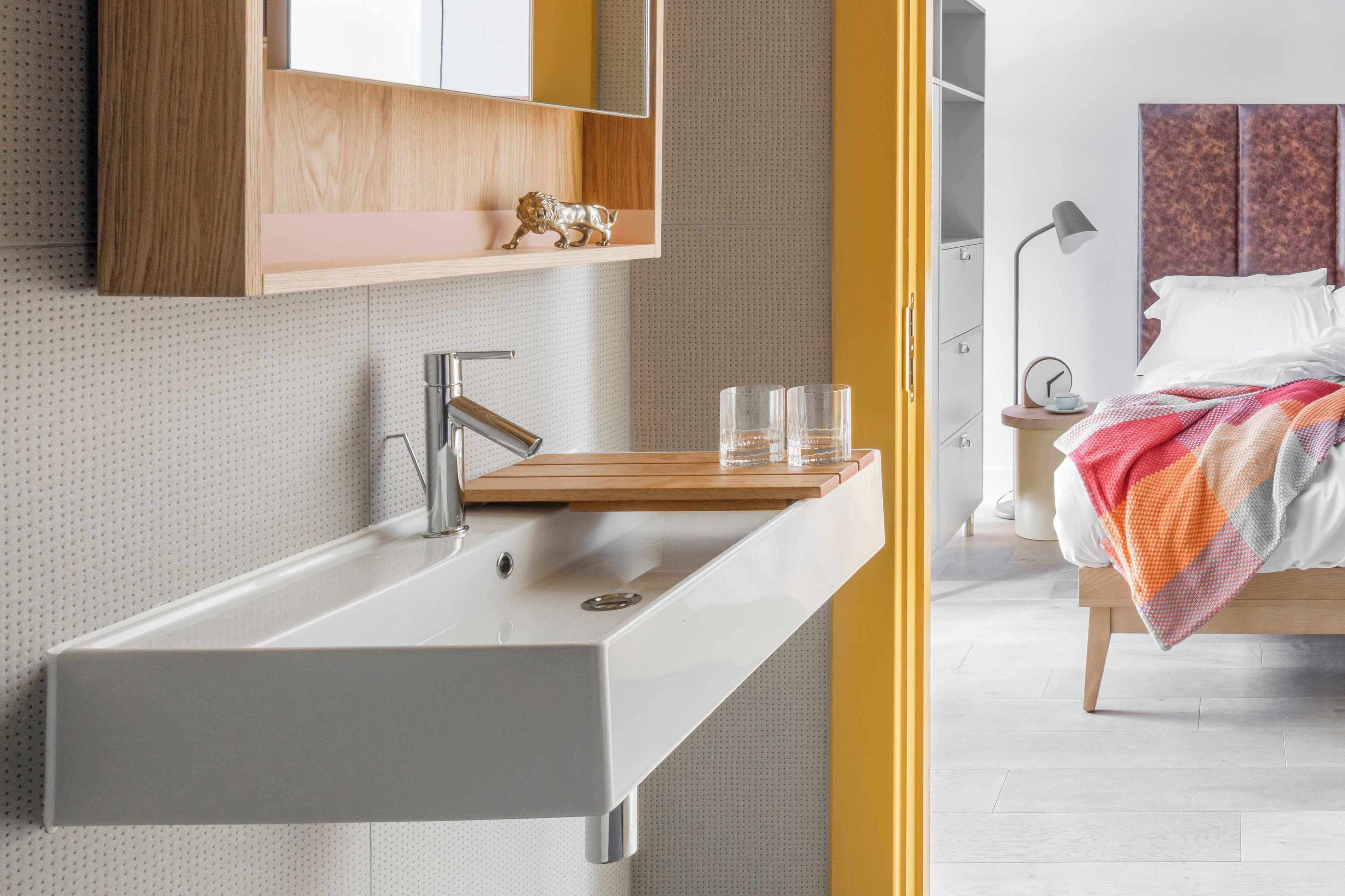 Une salle de bain moderne avec un lavabo intégré, une étagère en bois agrémentée d’un objet décoratif et une table de chevet avec une lampe et des lunettes.