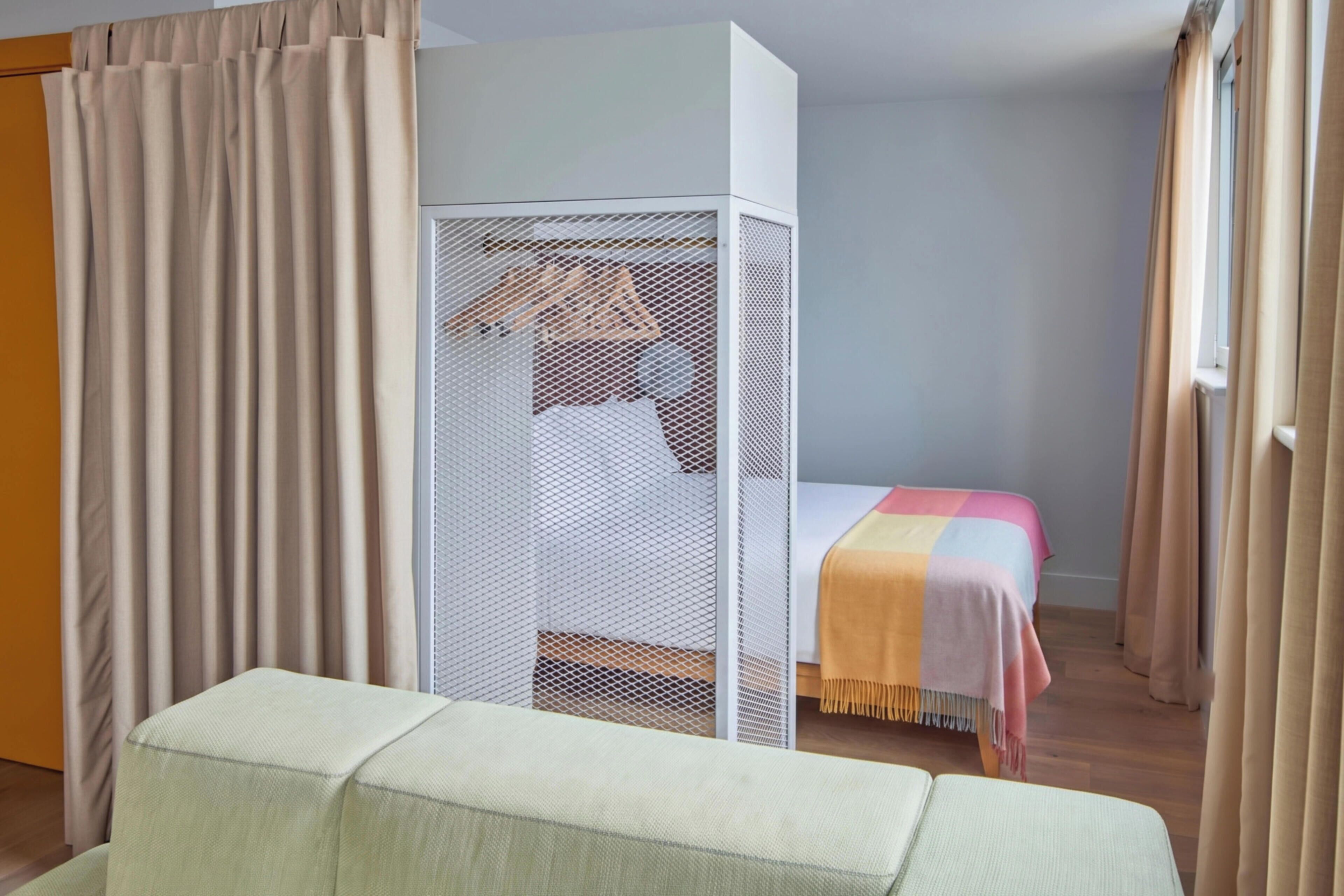 A room with a bed, a white partition, a hanging wardrobe, and a window with curtains.