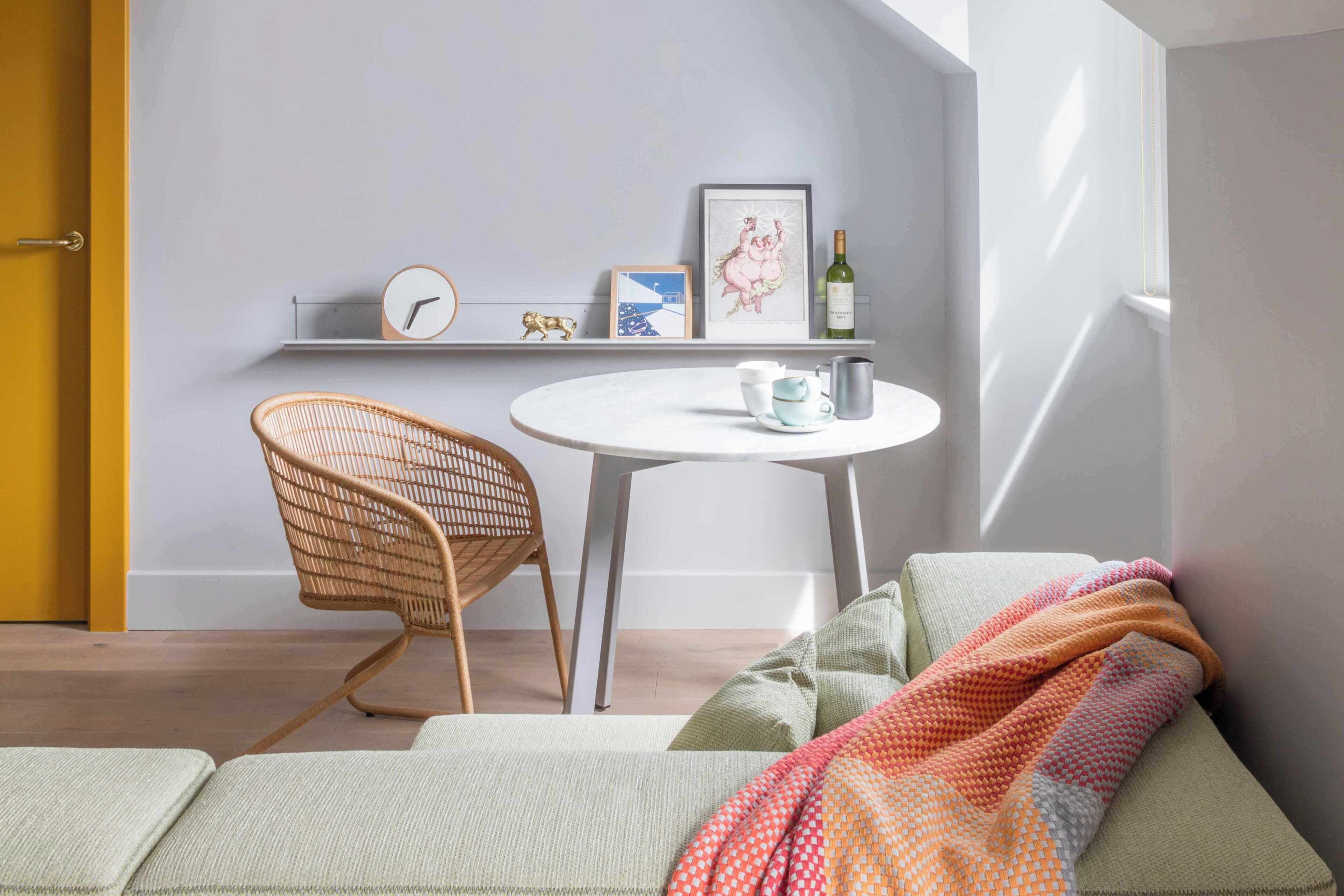 A room with a sofa, a wicker chair, a small round table with a clock, a framed picture, and a bottle.