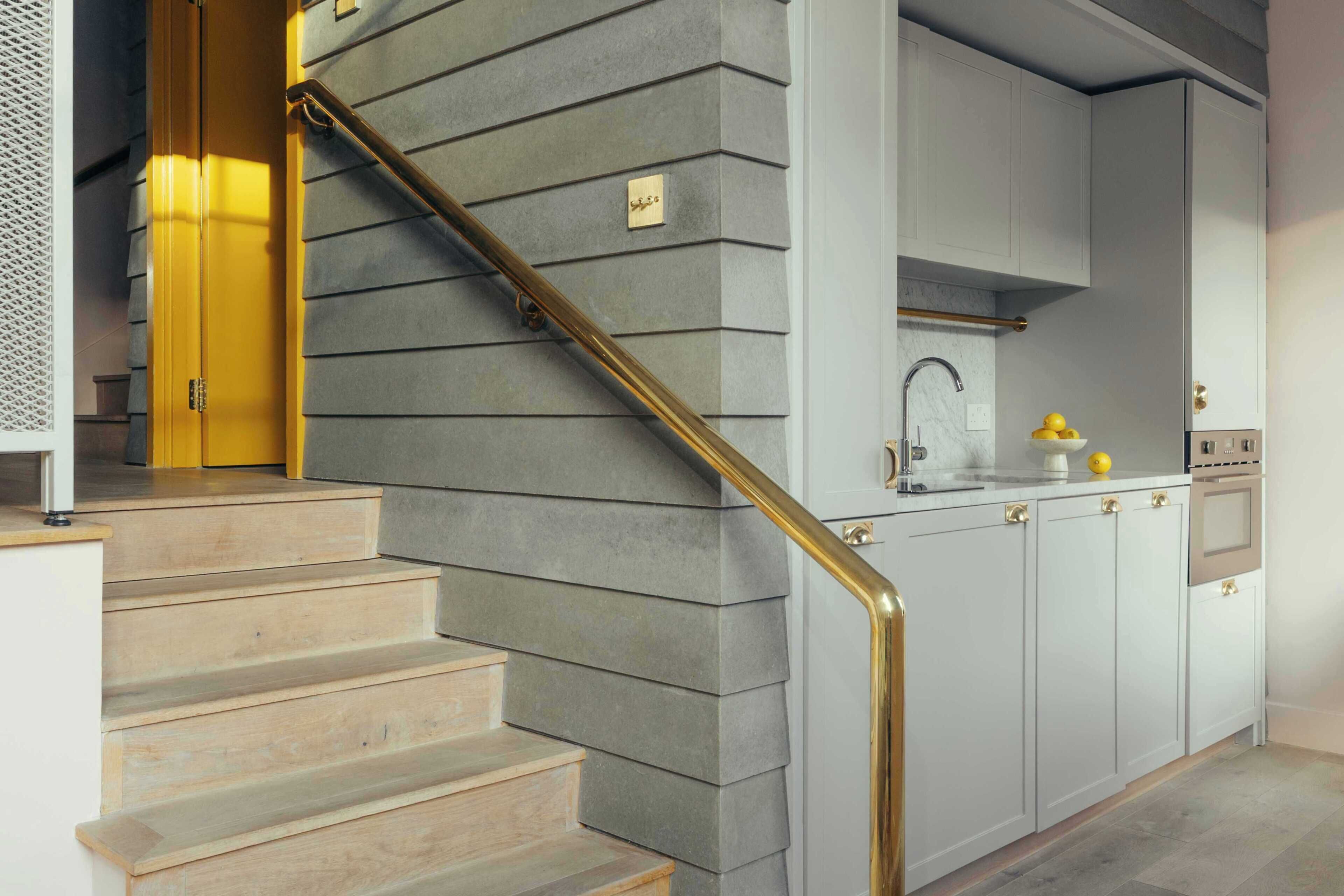 Un escalier doté d’une rampe dorée mène à une cuisine avec des meubles blancs et un évier en acier inoxydable.