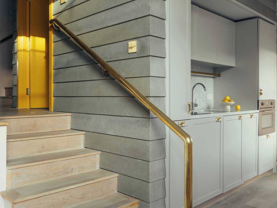 Un escalier doté d’une rampe dorée mène à une cuisine avec des meubles blancs et un évier en acier inoxydable.