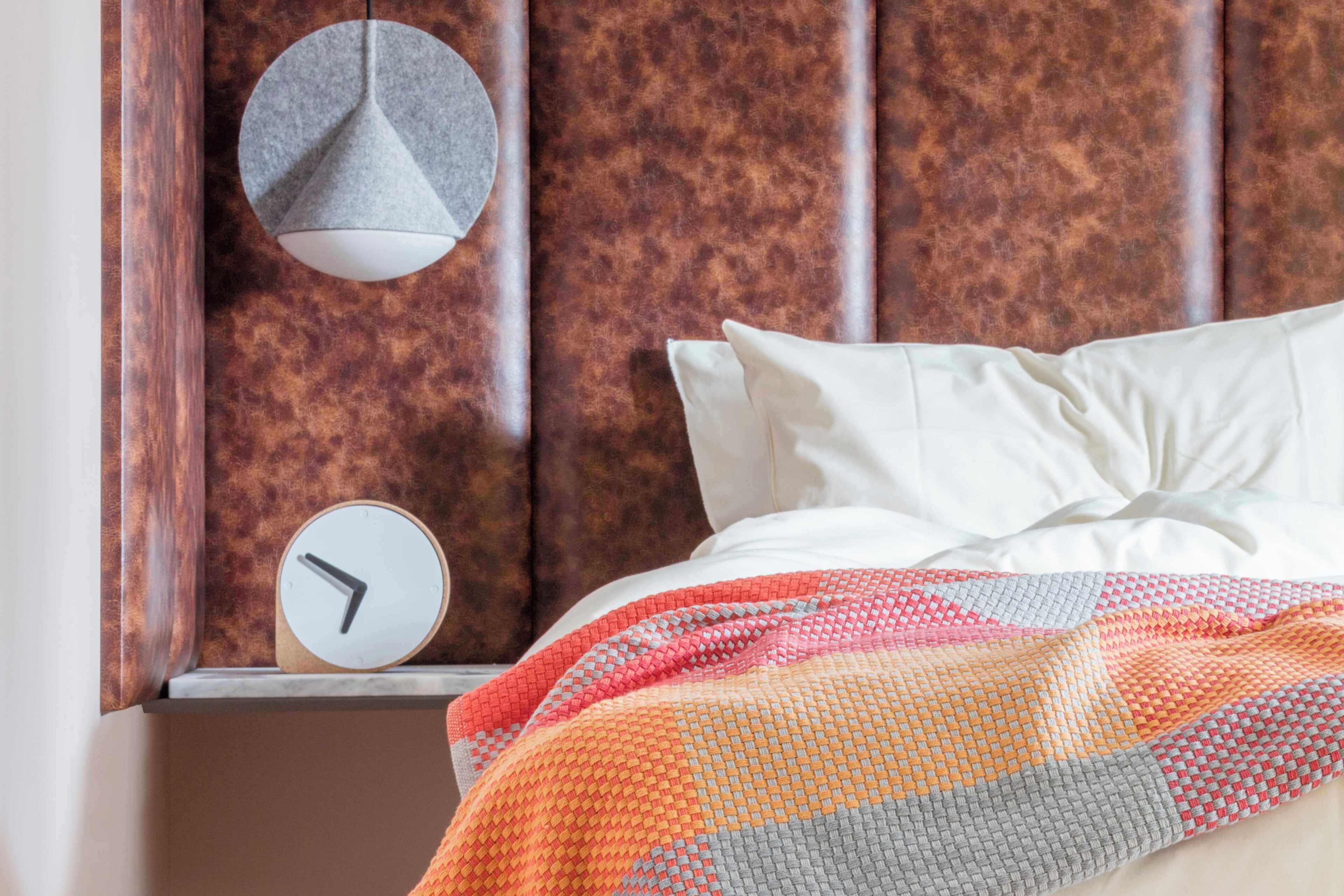 A bed with a colorful blanket, a round wall clock, and a pendant light.