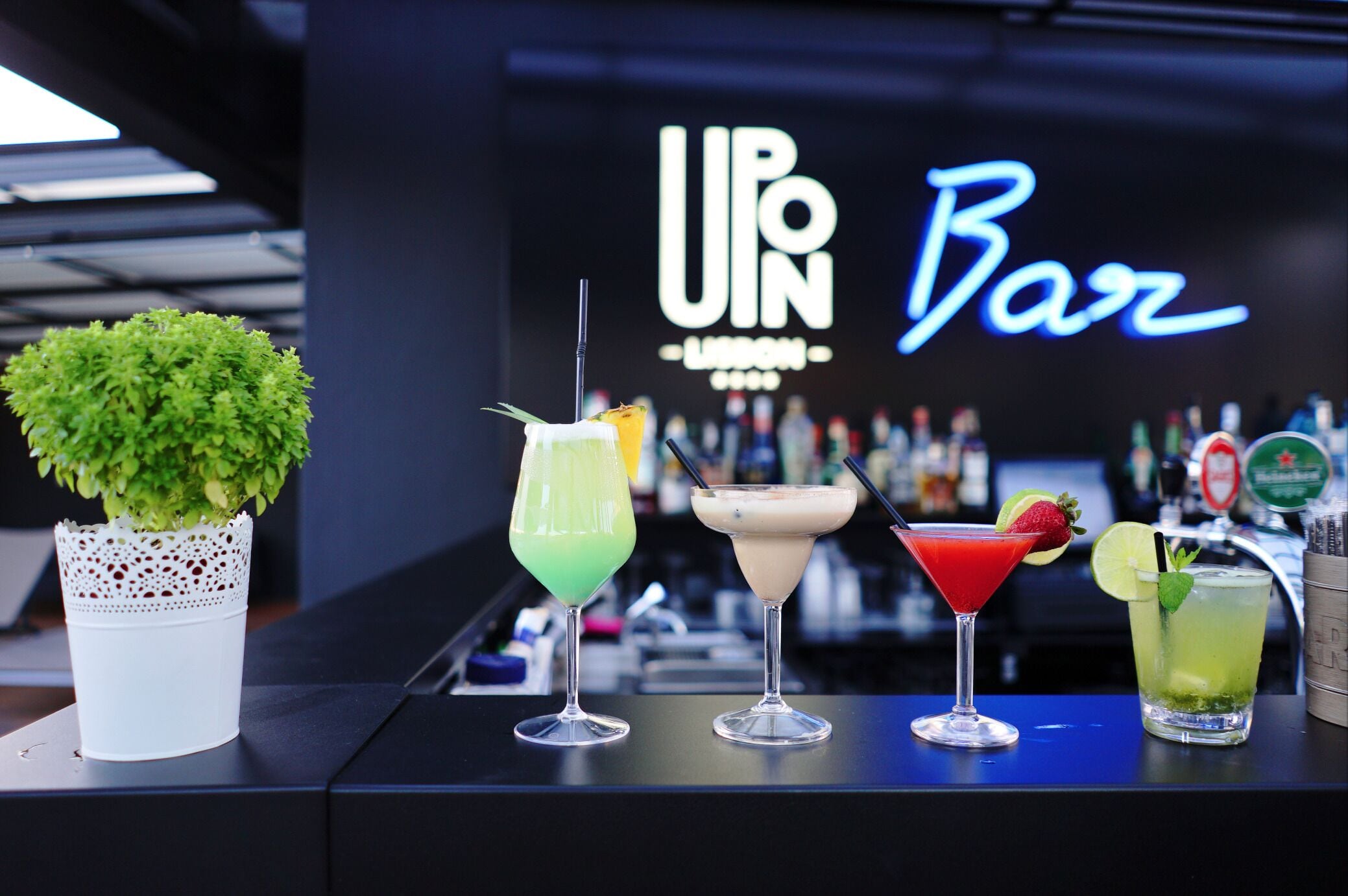 A bar counter with four cocktails and a potted plant. The background features a neon sign that reads "UPN Bar".