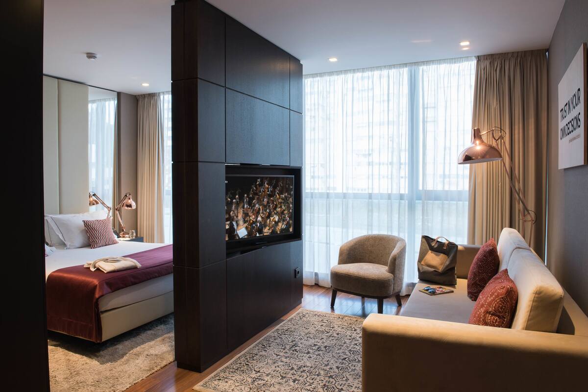 Quarto de hotel moderno com cama, sofá, televisão e uma grande janela com cortinas.