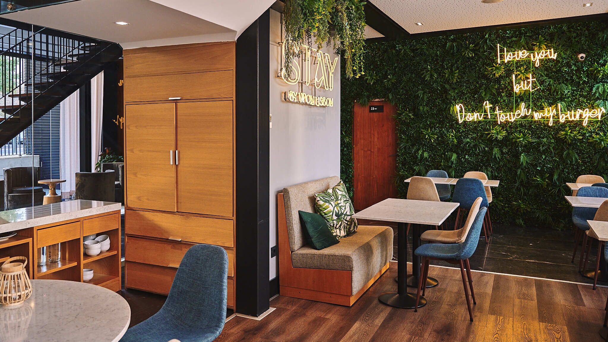 Un café moderno con mobiliario de madera, una pared verde con plantas y un letrero de neón.