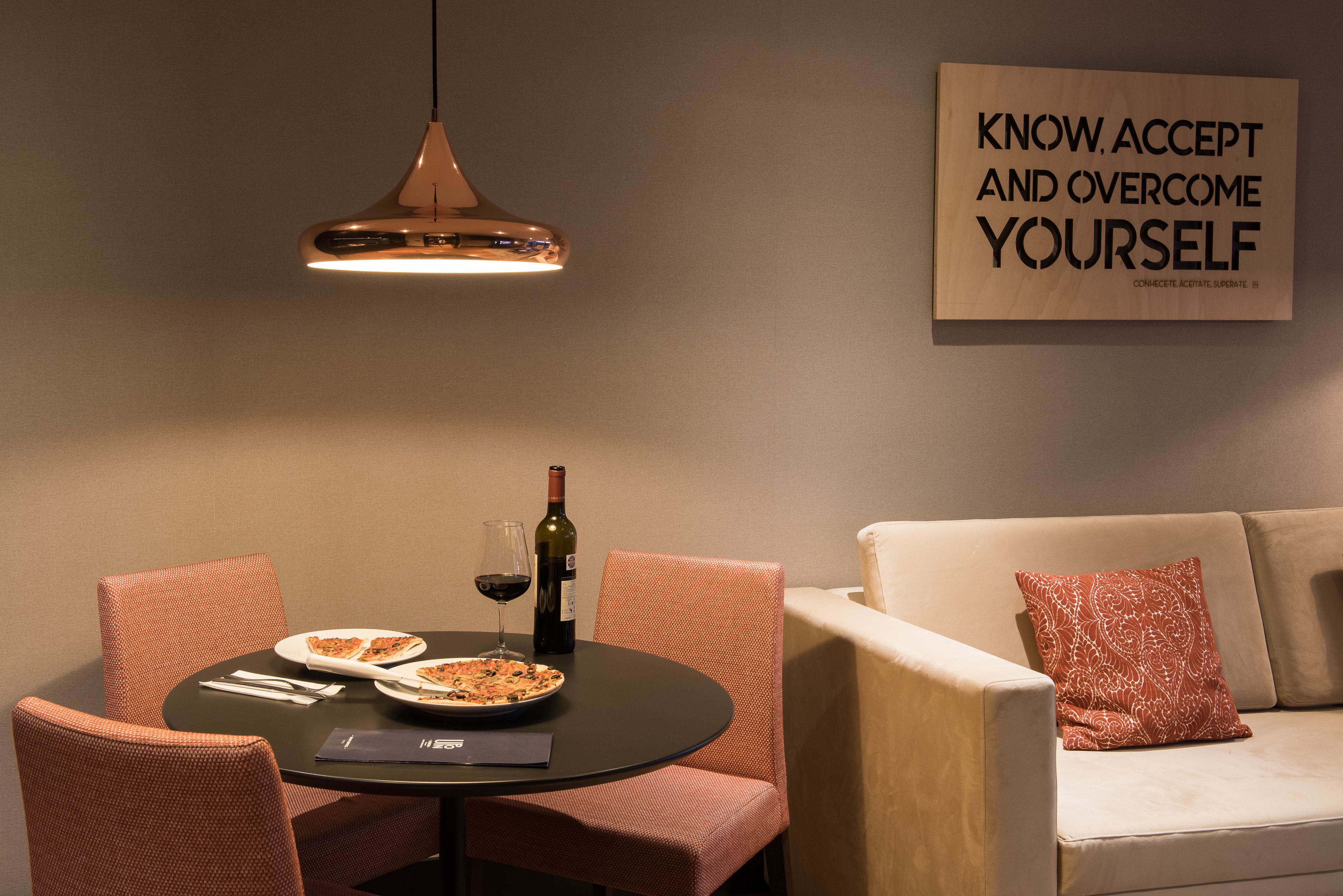A dining area with a round table, chairs, a bottle of wine, and a pizza. There is a sofa with a cushion and a motivational poster on the wall.