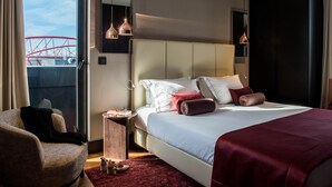 A modern hotel room with a large bed, a sitting area with a chair, a small side table, and a view of a roller coaster through the window.