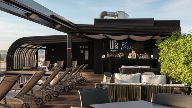 A rooftop bar with lounge chairs and tables, a bar area with a neon sign, and a view of the city.