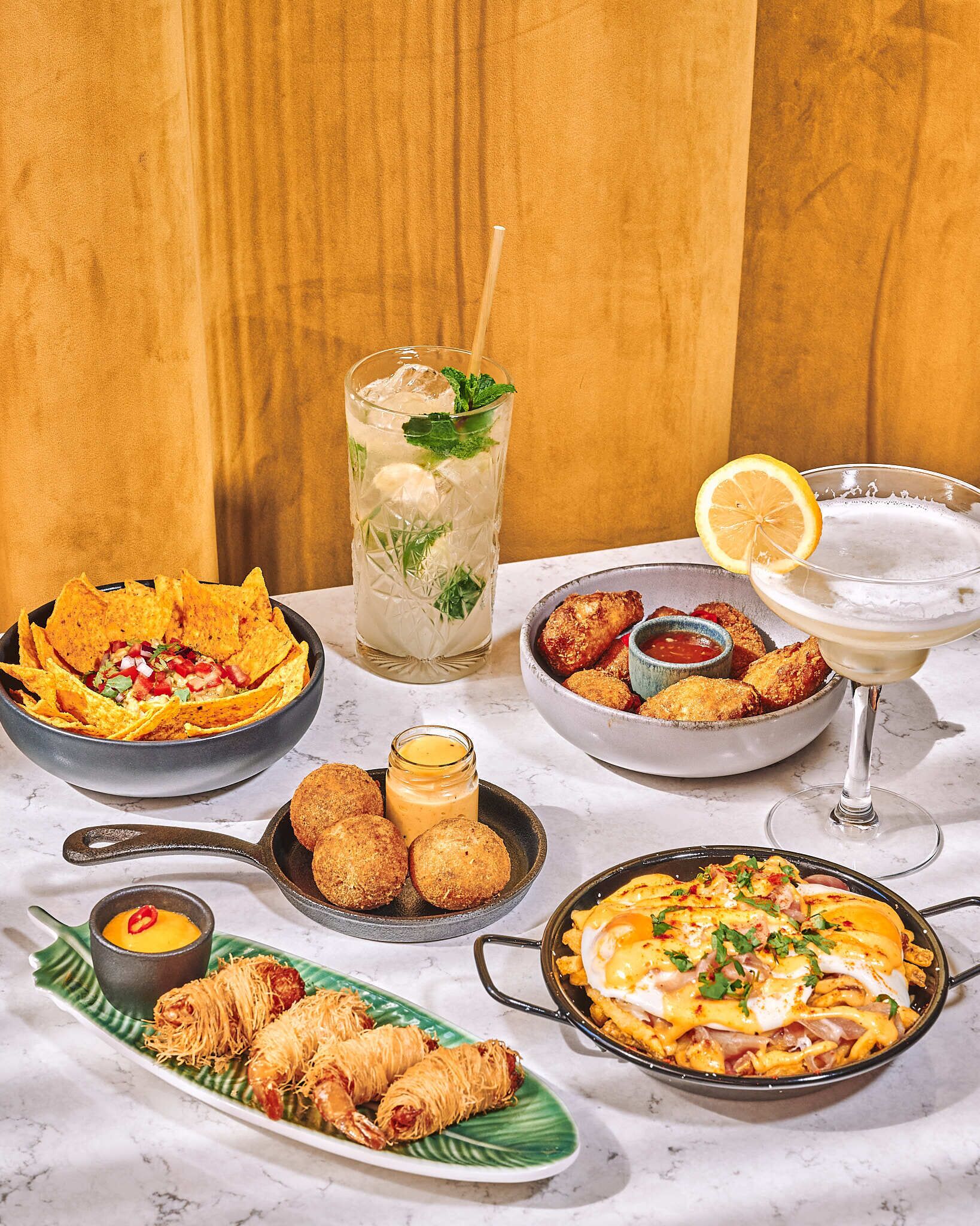 A variety of dishes including nachos, empanadas, and a pasta dish, along with drinks and condiments.