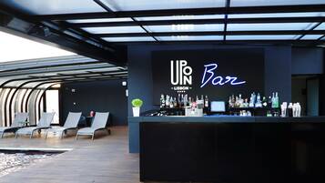 A bar area with a modern design, featuring a bar counter with a variety of bottles and a neon sign that reads "UPN Bar - Lisbon". There are lounge chairs and a small plant on the bar counter.
