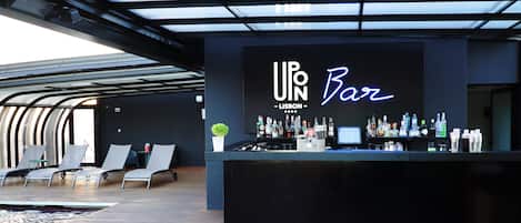 Un espace bar au design moderne, avec un comptoir bien garni de bouteilles et un néon indiquant « UPN Bar – Lisbonne ». On y trouve également des fauteuils confortables et une petite plante sur le comptoir.