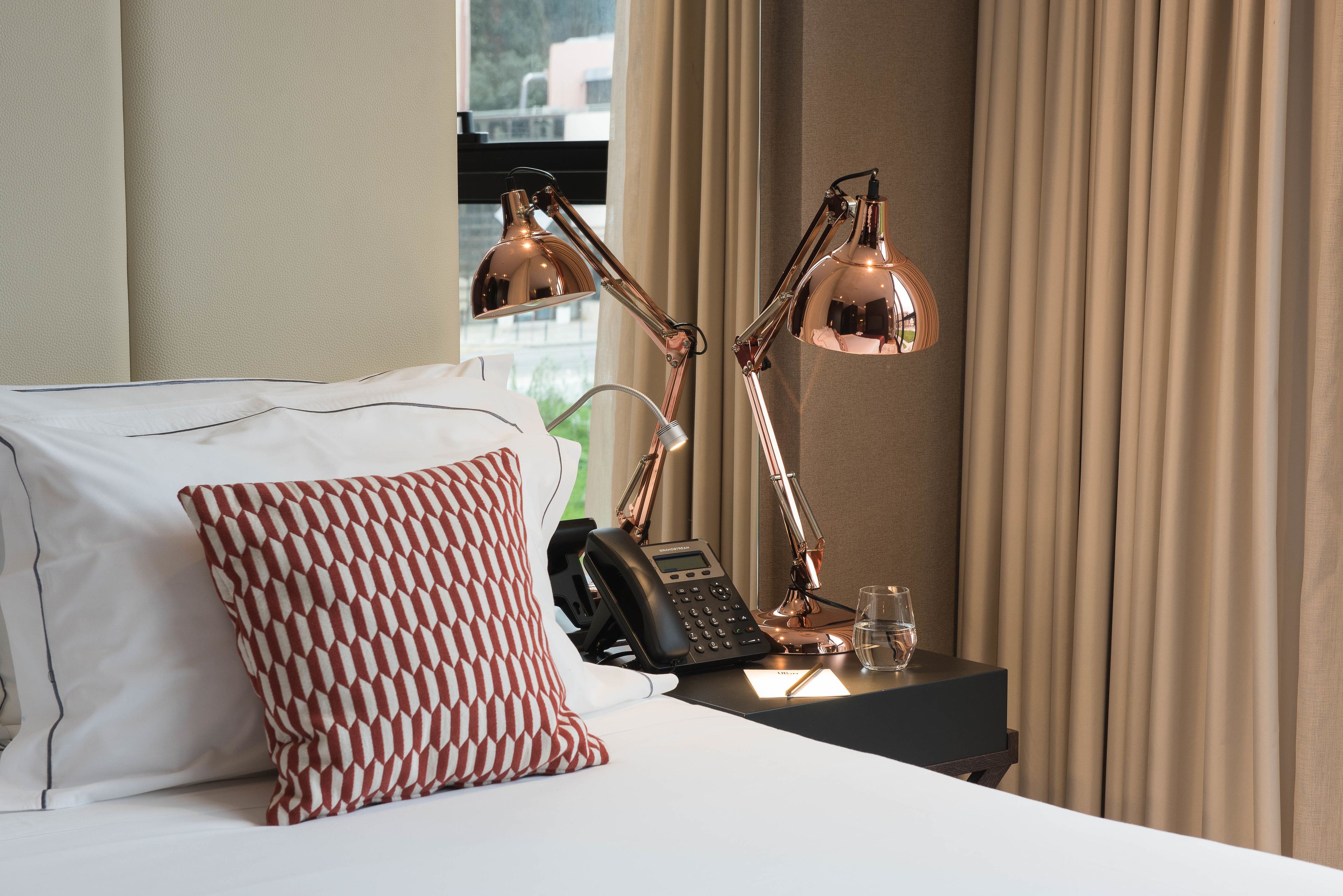 Habitación de hotel con cama, almohada estampada, escritorio con teléfono, lámpara y ventana con cortinas.