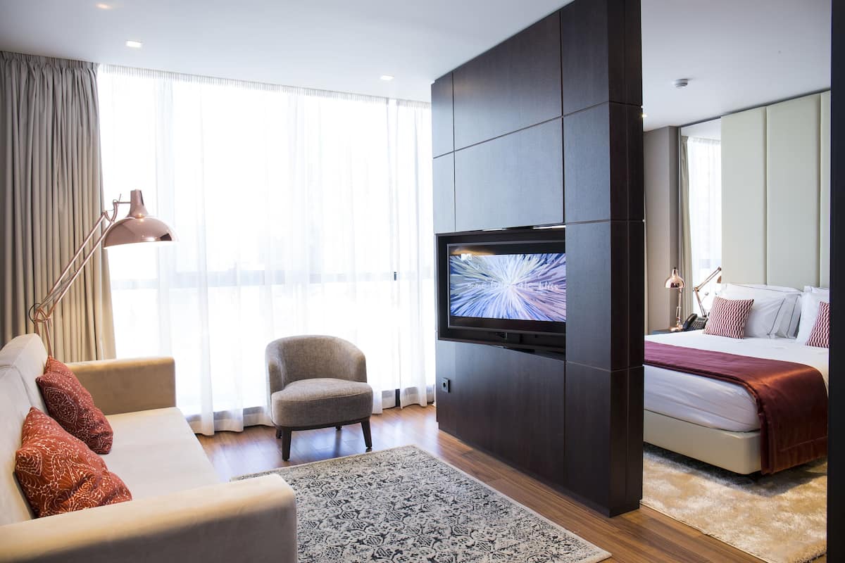 Quarto de hotel moderno com televisão de tela plana na parede, sofá com almofadas estampadas, uma poltrona pequena, cama com manta vermelha e uma janela grande com cortinas leves.