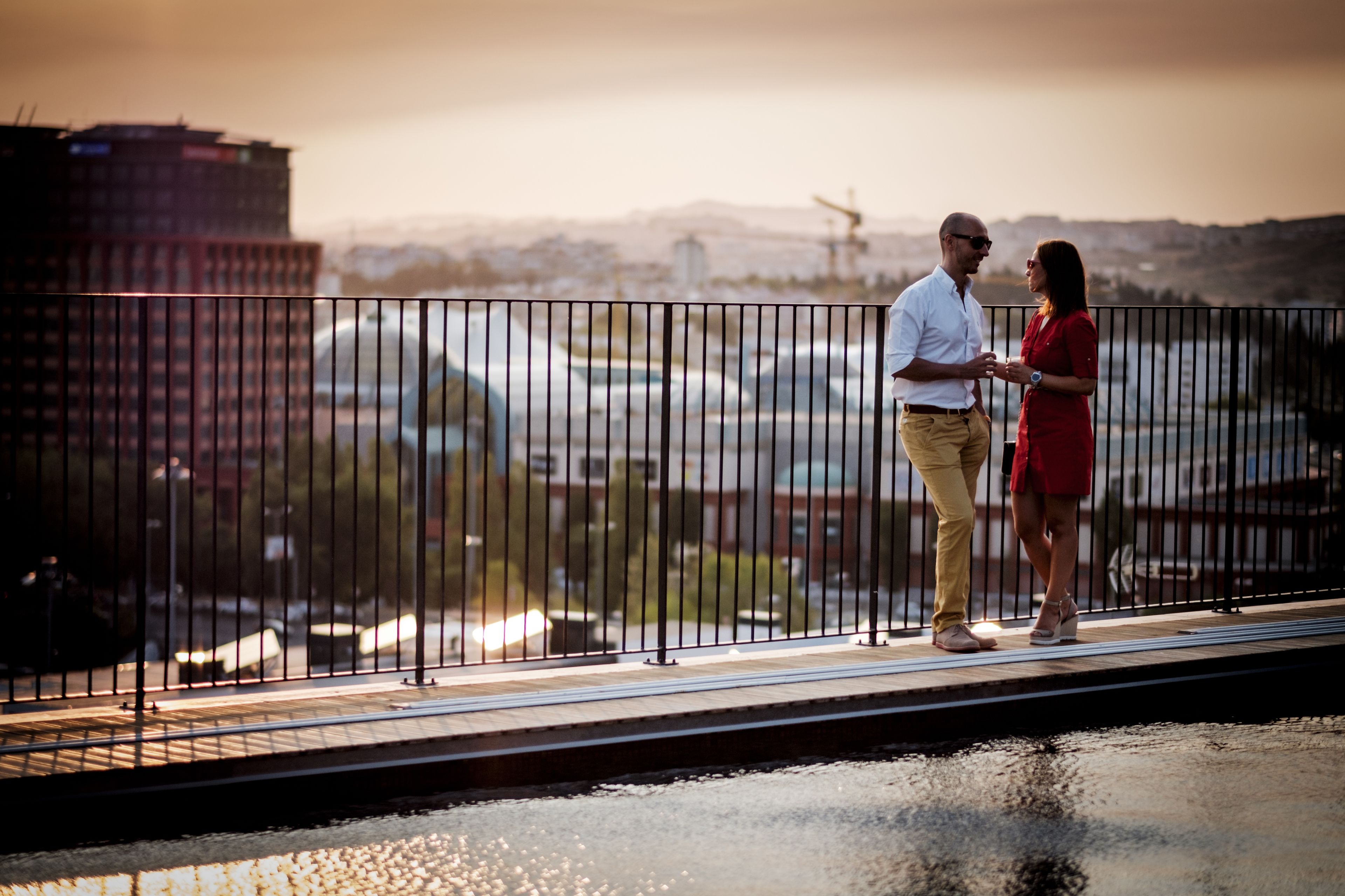 Um casal parado em uma ponte, com uma paisagem urbana ao fundo.