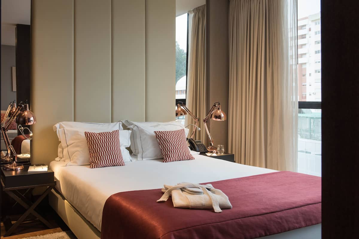Quarto de hotel com uma cama grande, luminárias de cabeceira, uma escrivaninha e vista para prédios pela janela.