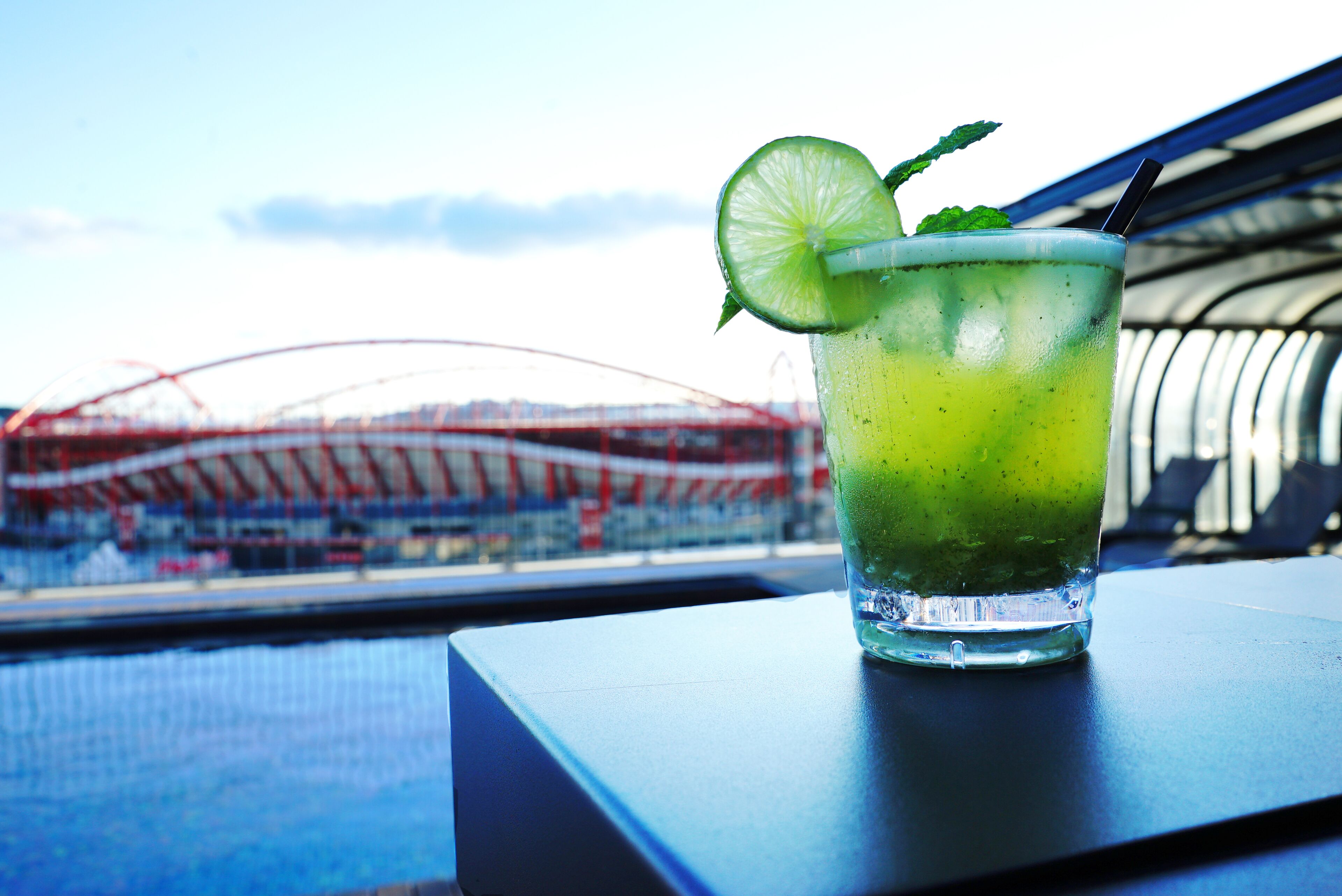 A glass of green drink with a lime slice and mint on a table with a view of a stadium in the background.