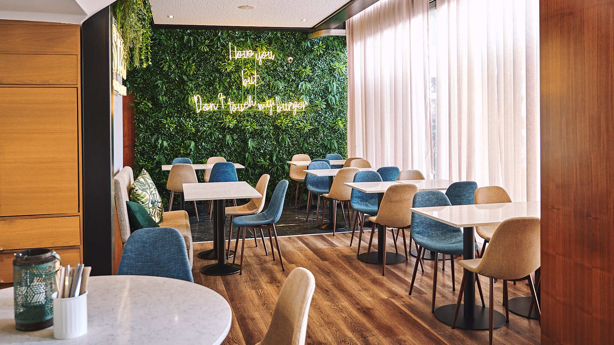 A modern restaurant interior with wooden flooring, a green wall with a neon sign, and blue and beige chairs around tables.