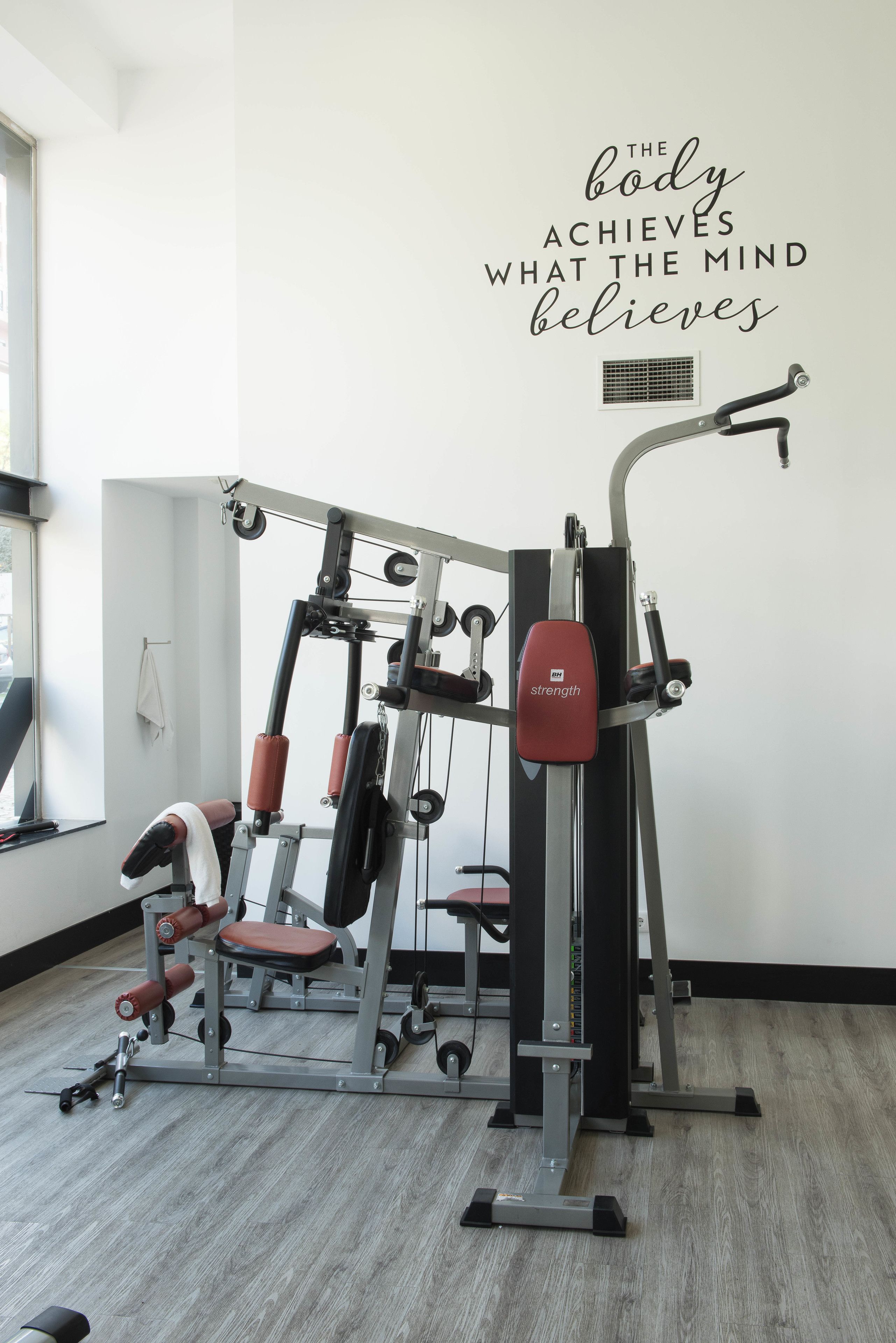 A gym with a wall-mounted motivational quote, a variety of exercise machines, and a window providing natural light.