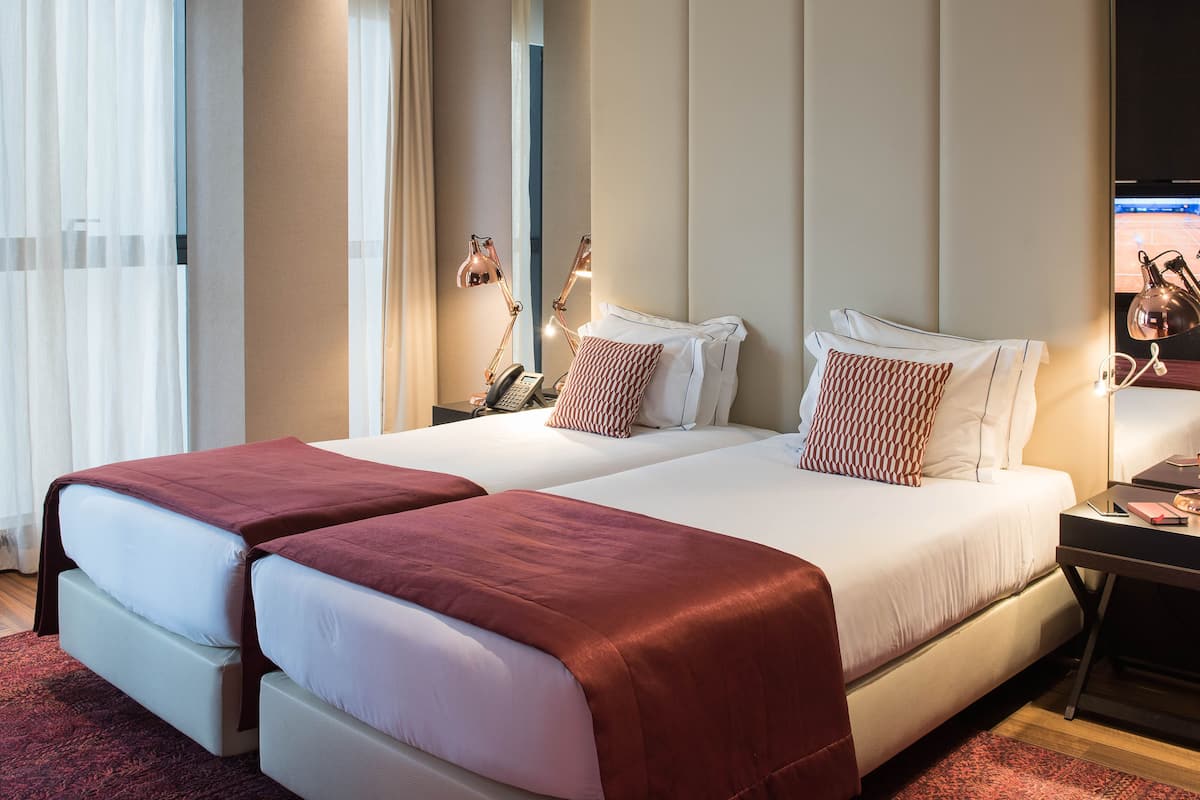 Cama de casal com duas almofadas, mesa de cabeceira com abajur e televisão à direita.