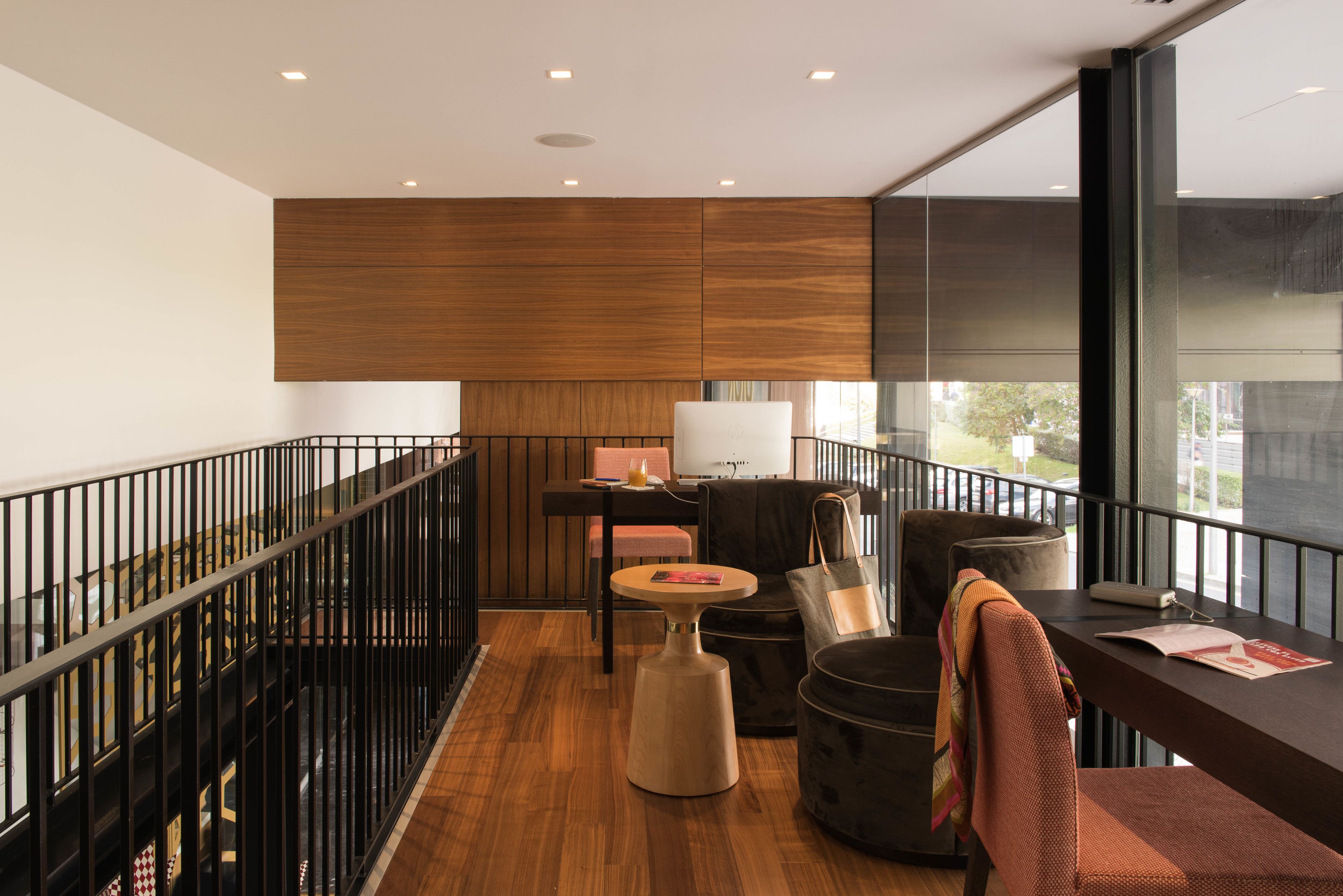 A modern hotel room with a wooden floor, a black railing, a round table, a sofa, and a large window offering a view of the outside.