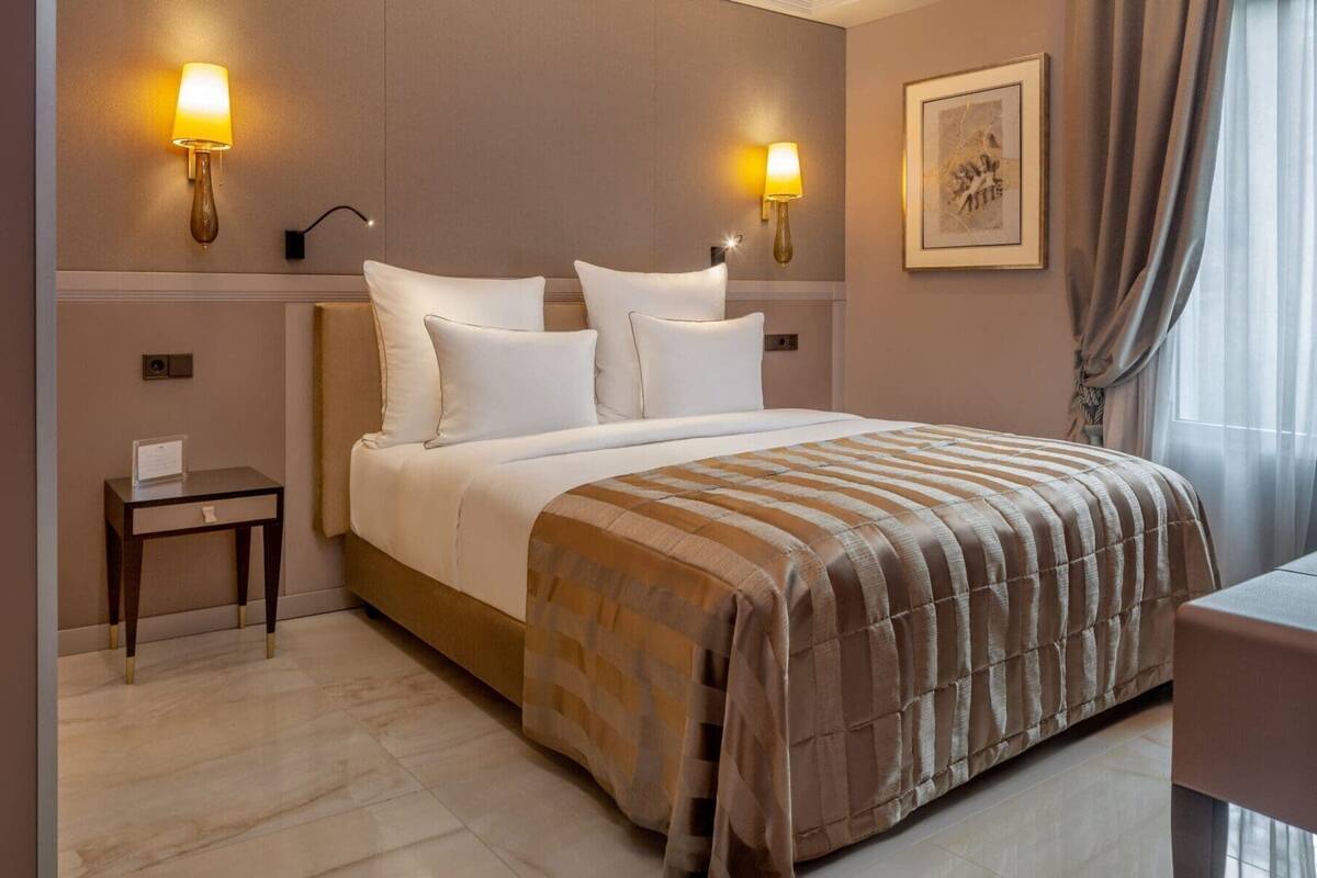 Une chambre d’hôtel avec un grand lit, deux lampes de chevet, une petite table, un tableau encadré au mur et une fenêtre avec des rideaux.