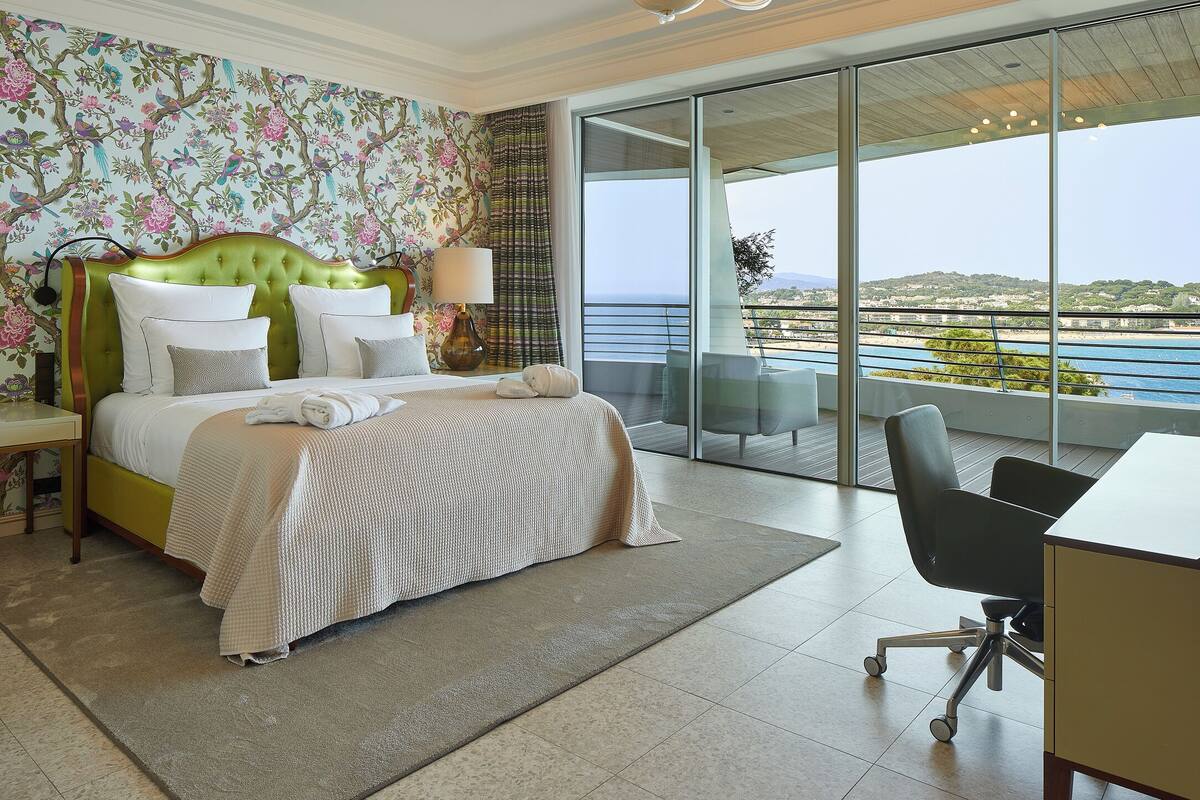 Une chambre d’hôtel avec un grand lit, un bureau et un balcon donnant sur la mer.