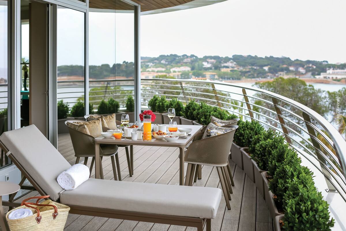Un espace extérieur lumineux, aménagé pour prendre les repas, avec une table dressée pour le petit-déjeuner, entourée de chaises en osier et d’un banc rembourré.