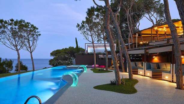 Un área de piscina moderna al aire libre, con una piscina azul cristalina, borde de agua infinita de cristal y un espacio de descanso con vistas al mar.