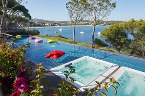 Une piscine à débordement offrant une vue sur la mer et les bateaux, entourée d’arbres et de chaises longues aux couleurs vives.