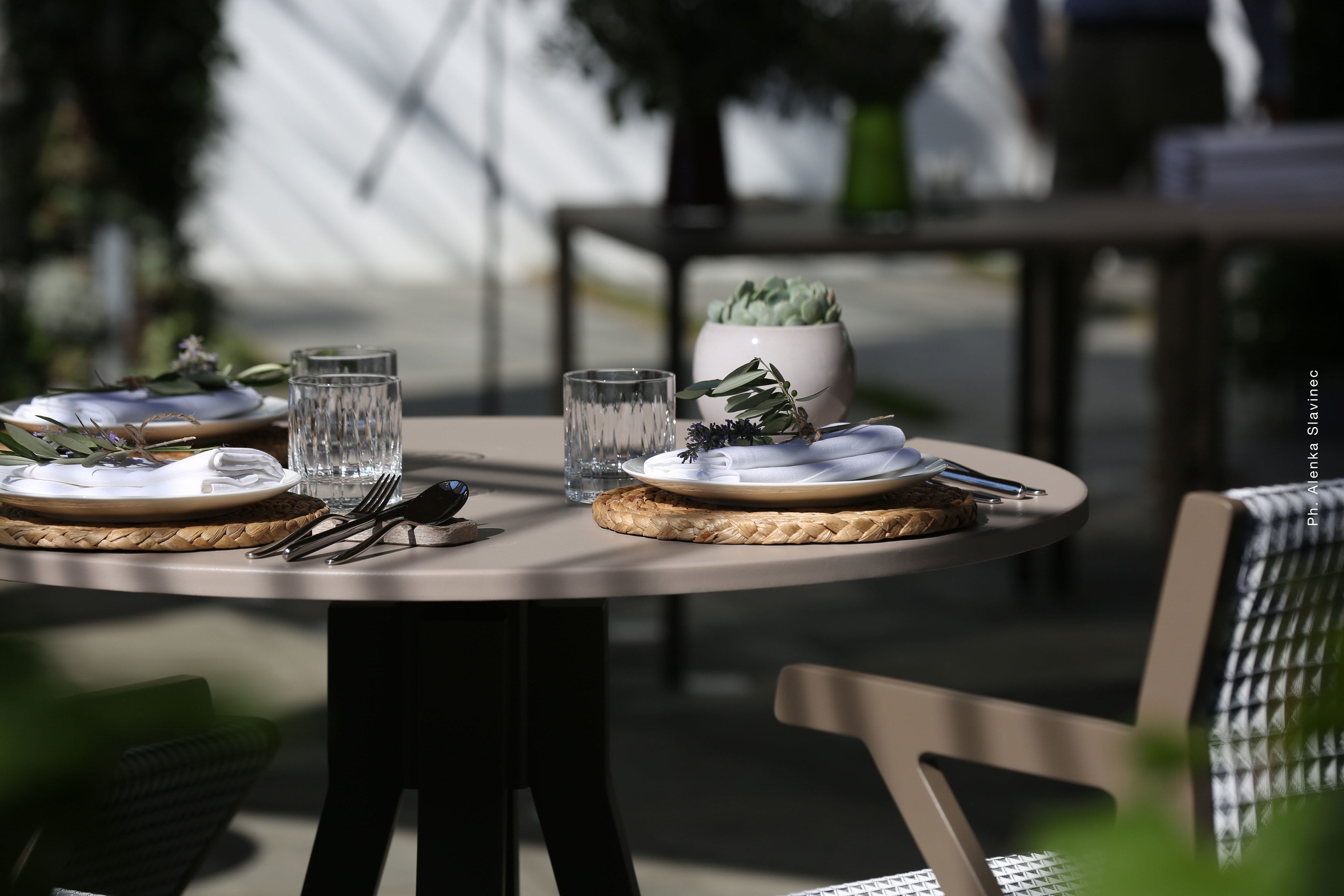 Mesa redonda para comer servida con platos, cubiertos y vasos, con un mantel tejido y una planta en maceta.