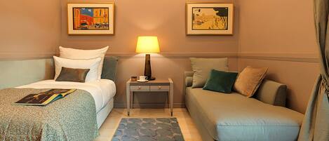 A hotel room with a bed, a sofa, a nightstand with a lamp, and two framed pictures on the wall.