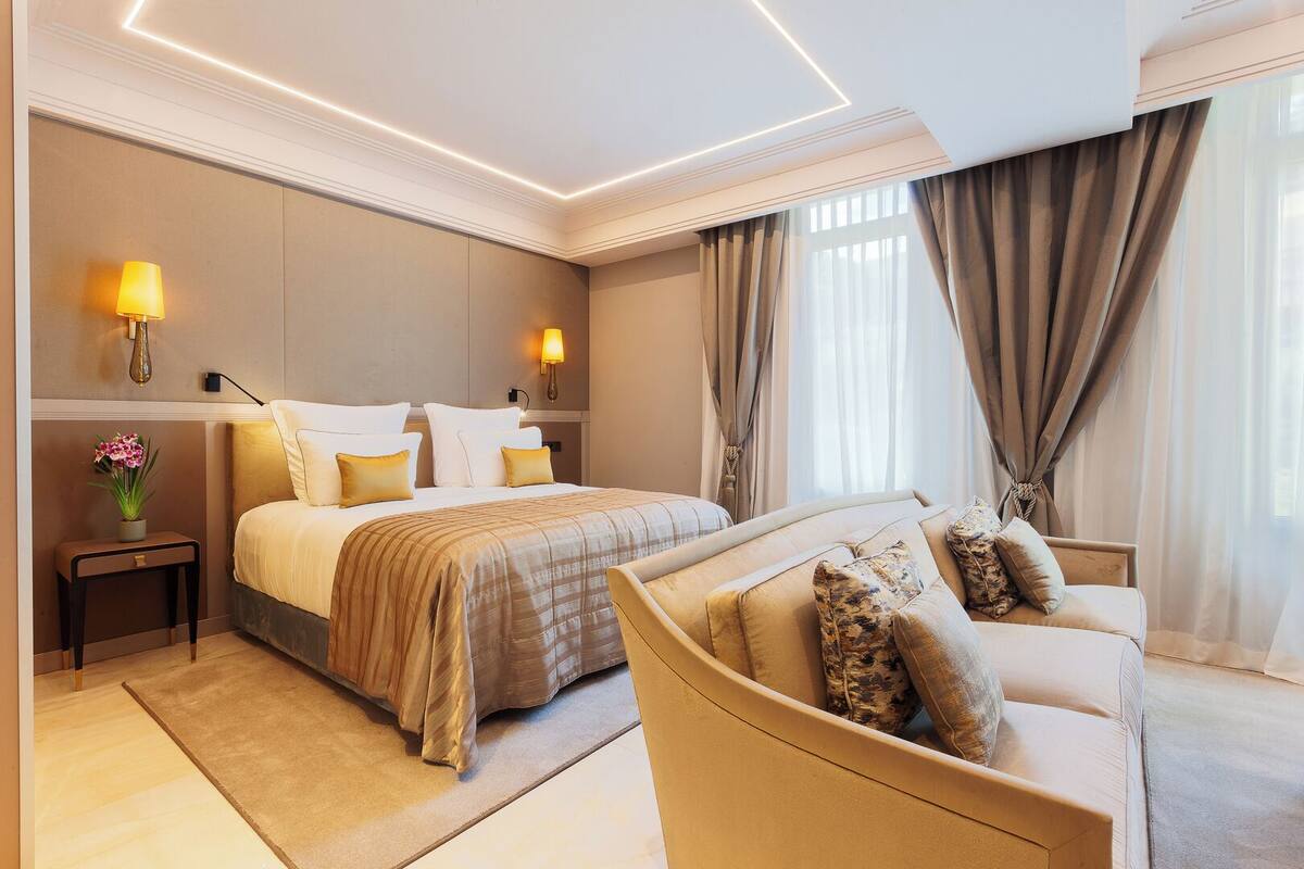 Une chambre d’hôtel avec un lit, un canapé, une table de chevet avec une lampe et une fenêtre avec des rideaux.