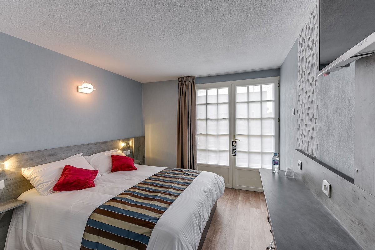 Ein modernes Hotelzimmer mit einem großen Bett, einem Nachttisch, einem wandmontierten Fernseher und einem Fenster mit Vorhängen.
