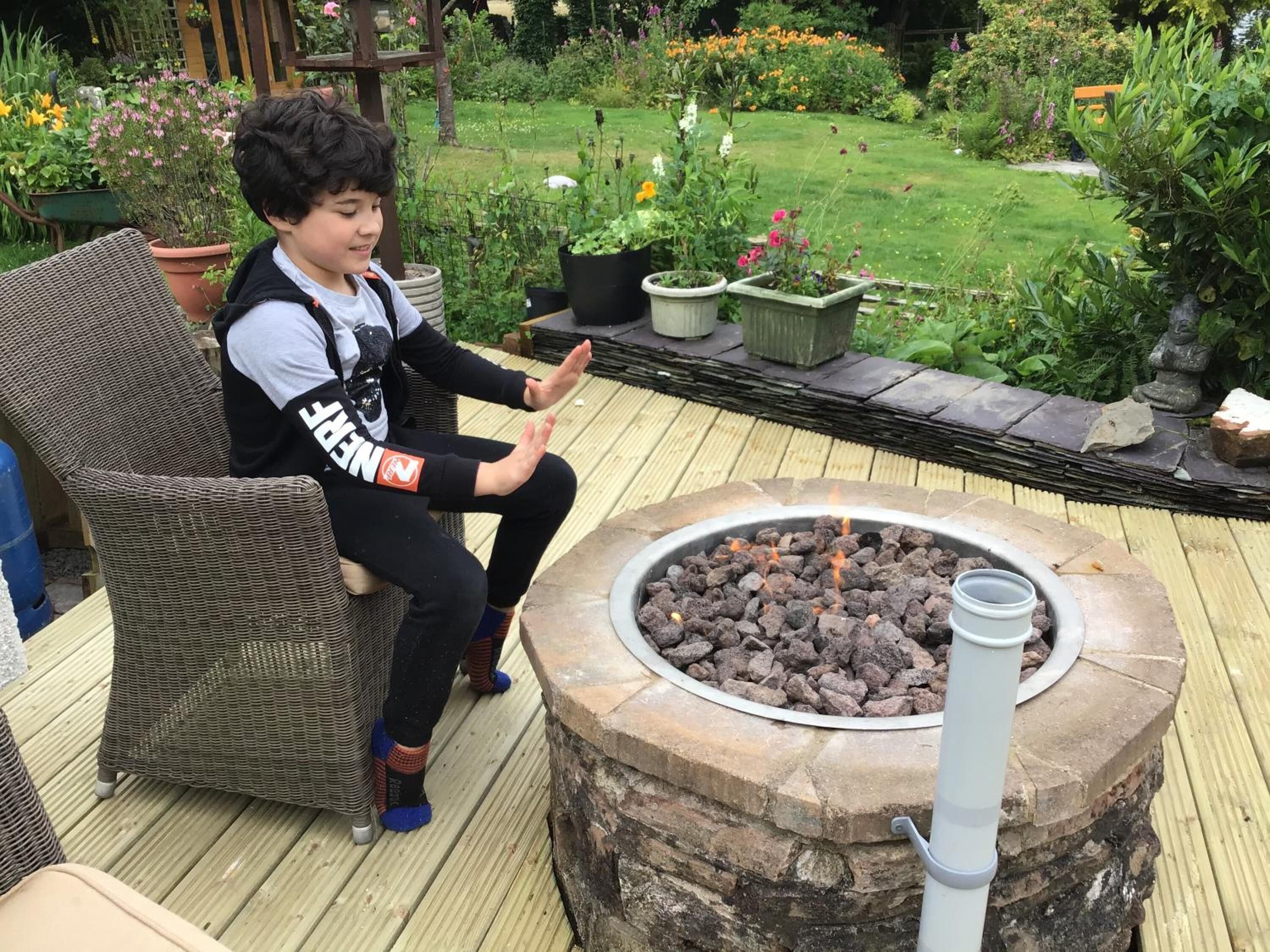 A child sitting on a wicker chair on a wooden deck with a stone fire pit in the center.