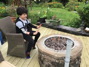 A child sitting on a wicker chair on a wooden deck with a stone fire pit in the center.