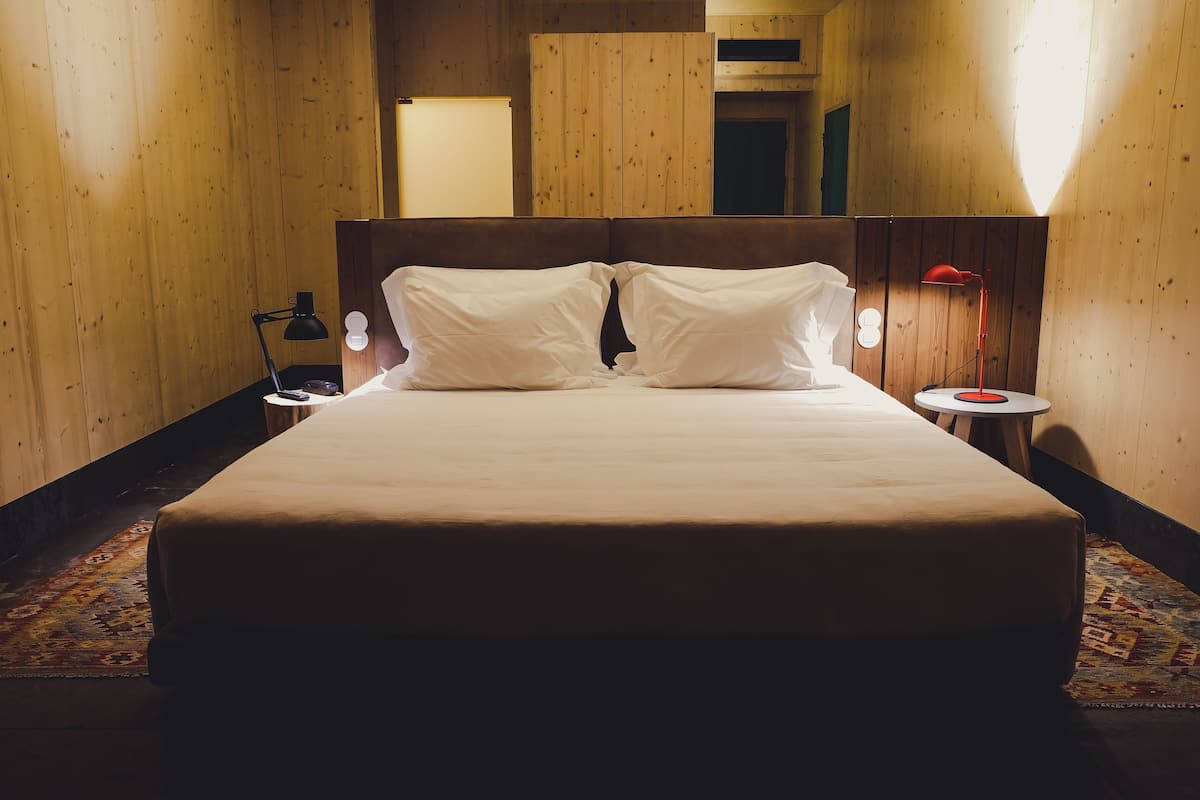 Un lit bien fait dans une pièce en bois, avec une table de chevet, une lampe et une petite table sur laquelle se trouve un objet rouge.