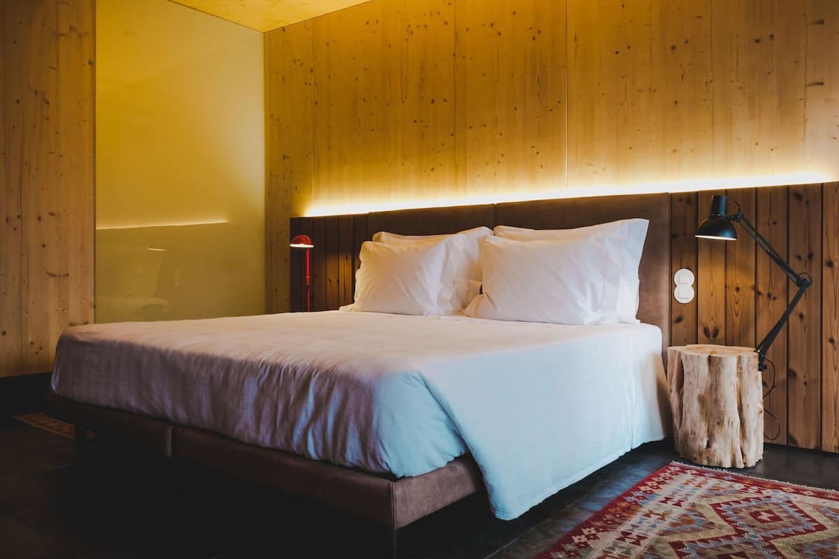 Une chambre à coucher avec une tête de lit en bois, un lit recouvert de draps blancs, une table de chevet avec une lampe et un tronc d’arbre servant de table d’appoint.