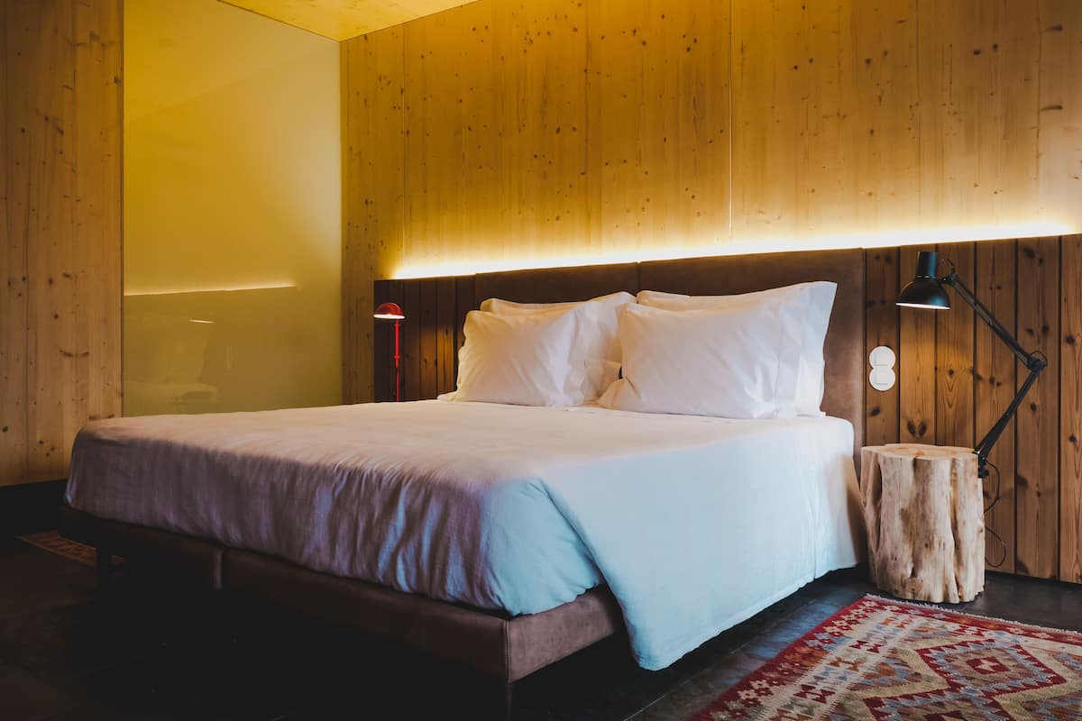 Une chambre à coucher avec une tête de lit en bois, un lit recouvert de draps blancs, une lampe de chevet et un tronc d’arbre servant de table de chevet, placé à côté du lit.