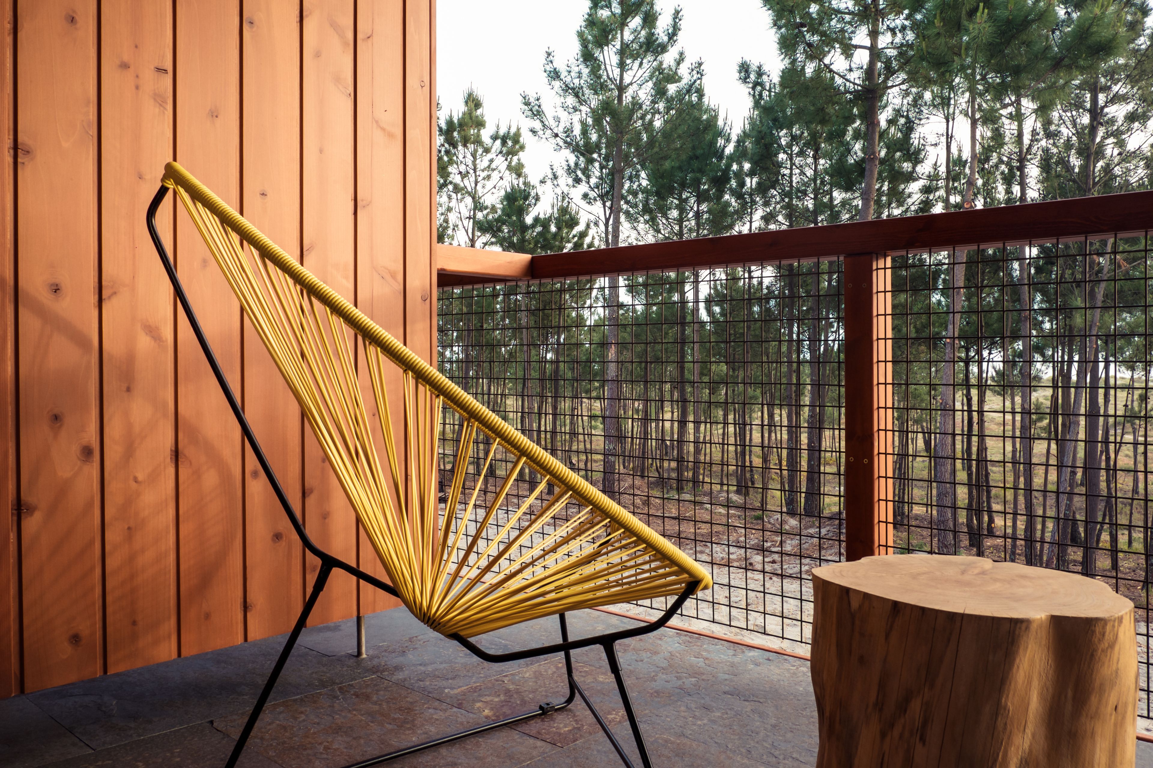 Une chaise jaune avec une structure noire, placée sur un balcon doté d’un mur en bois et d’une rambarde en métal.
