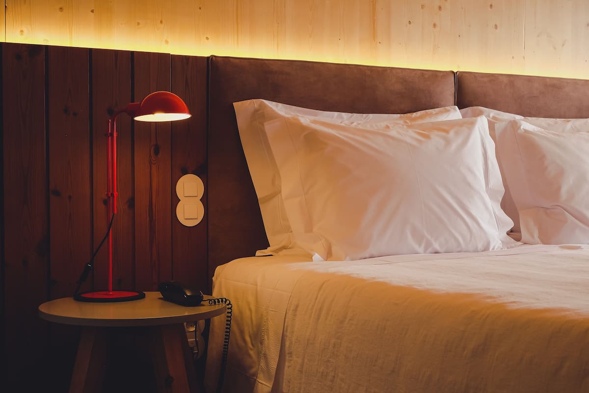 Un lit bien fait, avec du linge de lit blanc, une lampe rouge posée sur une petite table à côté, et un mur en bois en arrière-plan.