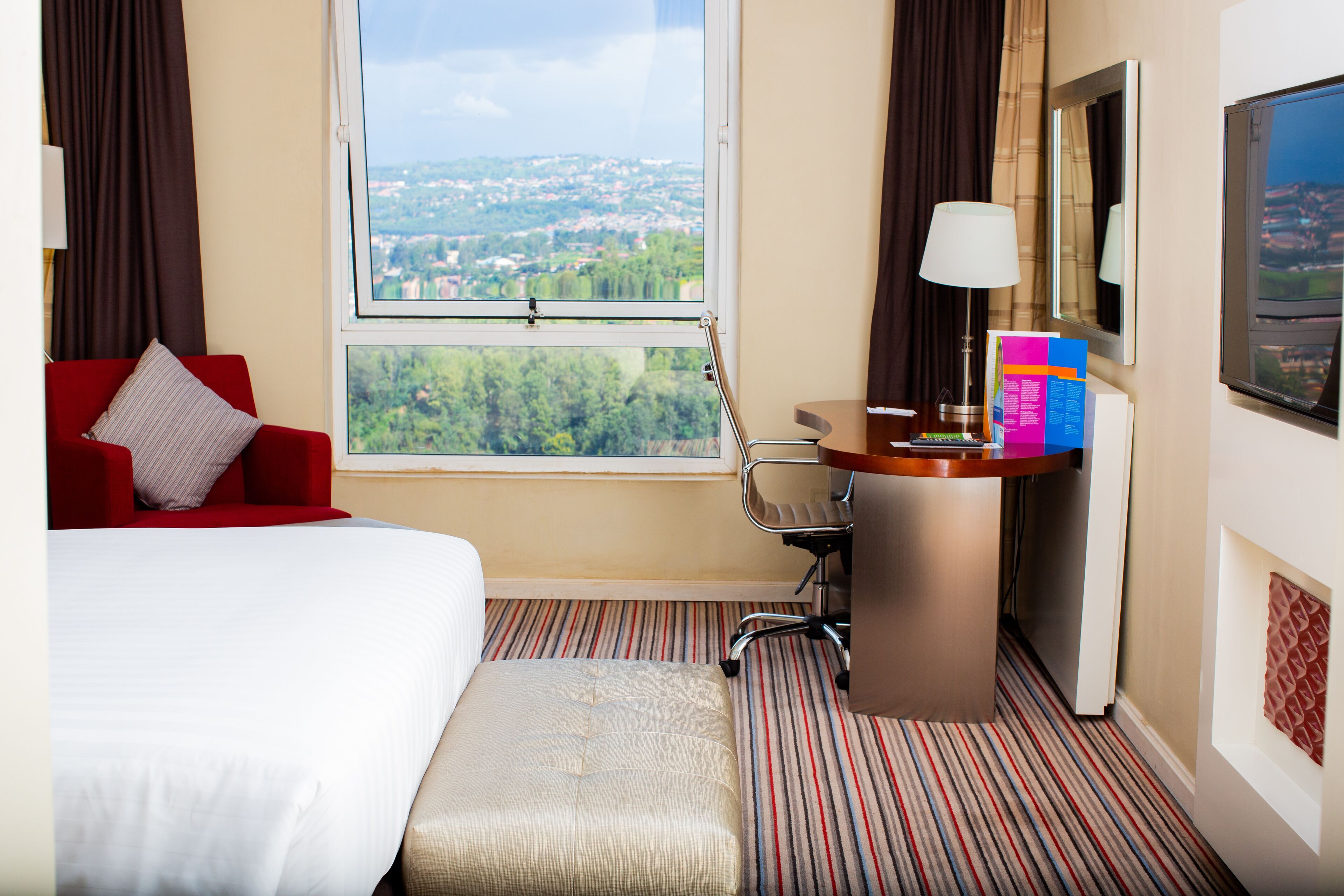 Ein Hotelzimmer mit einem Bett, einem roten Sessel, einem Schreibtisch mit Lampe, einem Stuhl und Blick auf eine Stadtansicht durch ein Fenster.