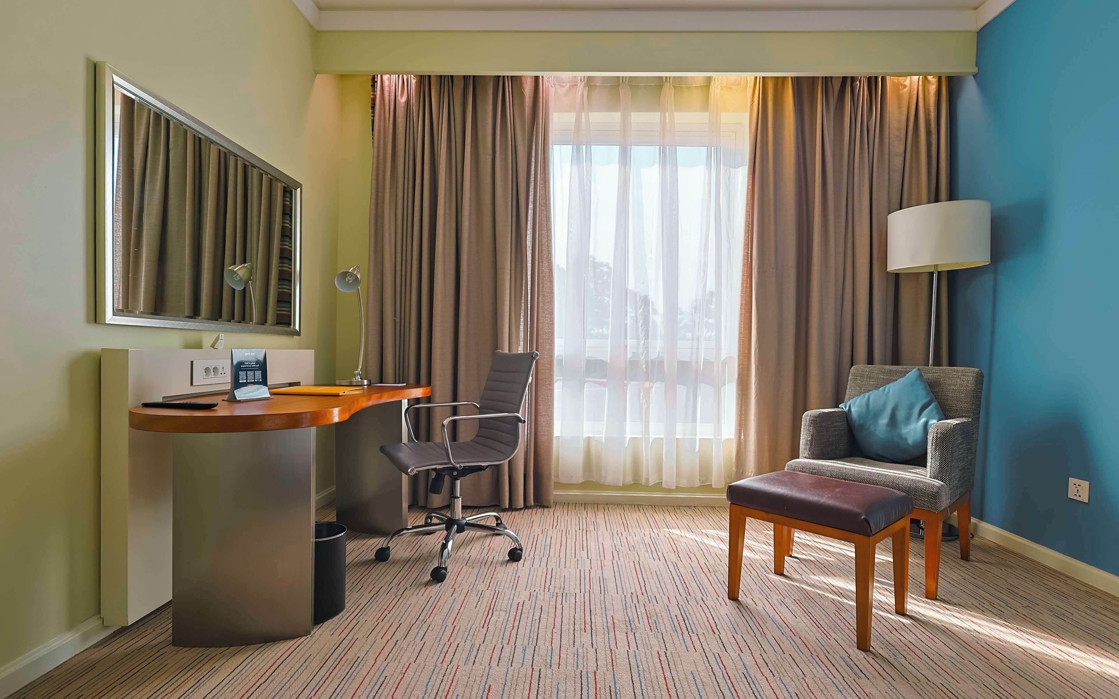 A hotel room with a desk, a chair, a sofa, a lamp, and a view of the outdoors.