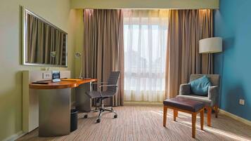 A hotel room with a desk, a chair, a sofa, a lamp, and a view of the outdoors.