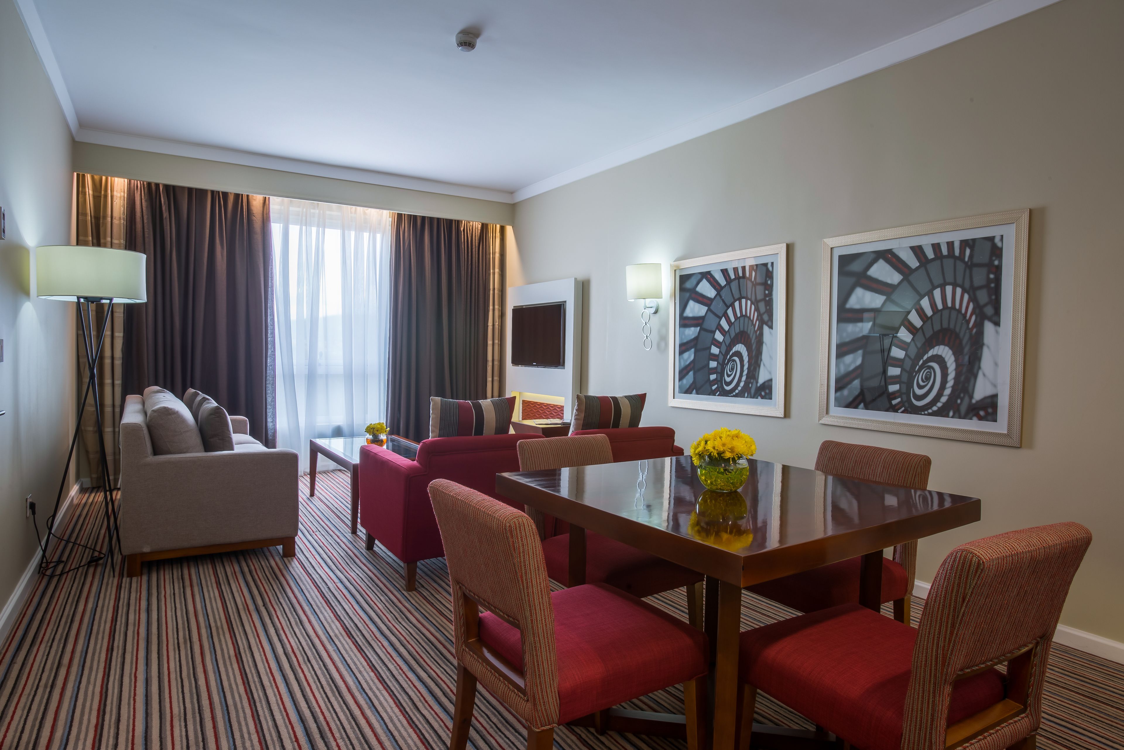 A hotel room with a striped carpet, a sofa, a dining table with chairs, a TV, and two framed pictures on the wall.
