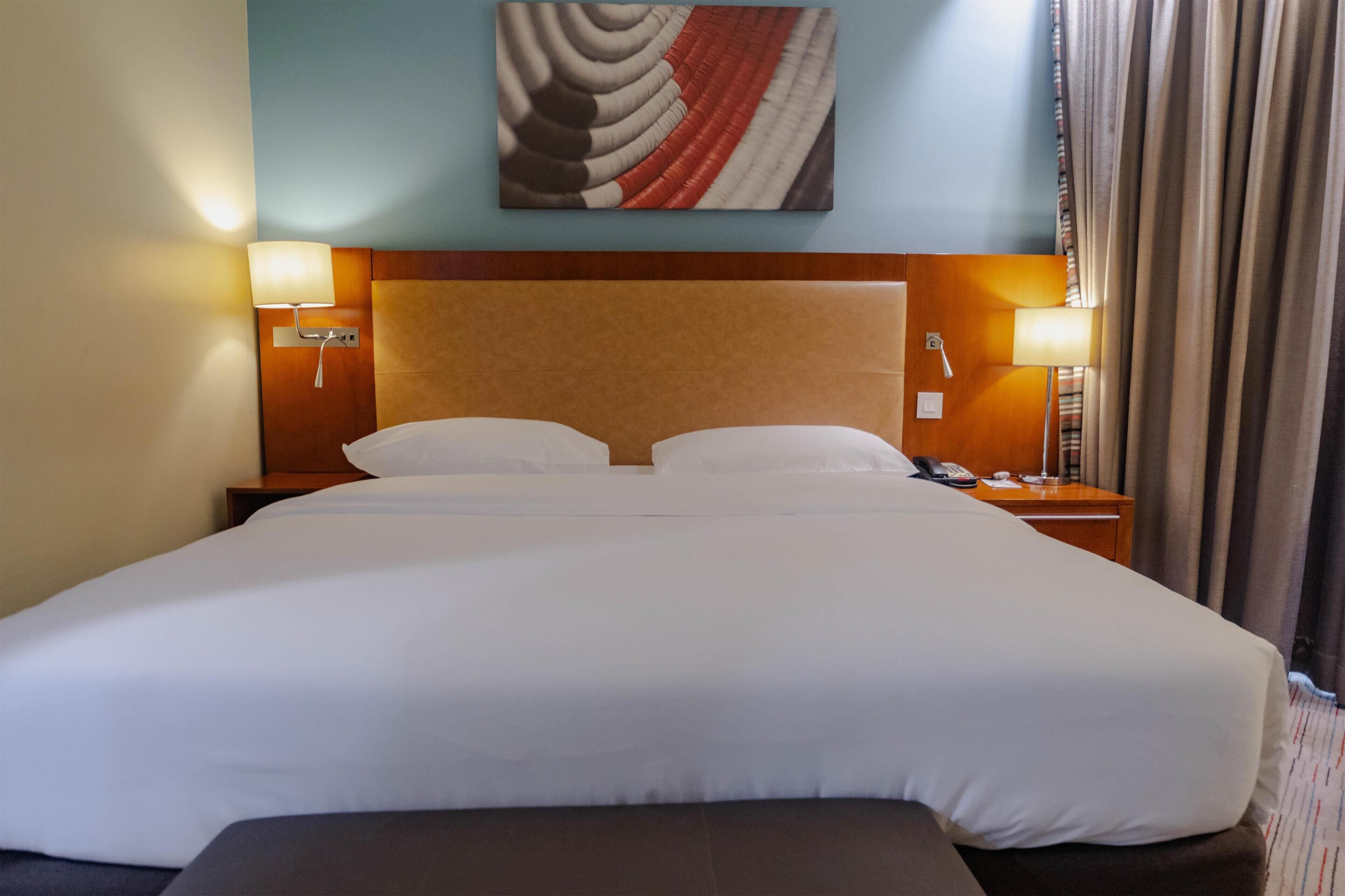 A hotel room with a large bed, two bedside lamps, a nightstand with a phone, and a painting on the wall.