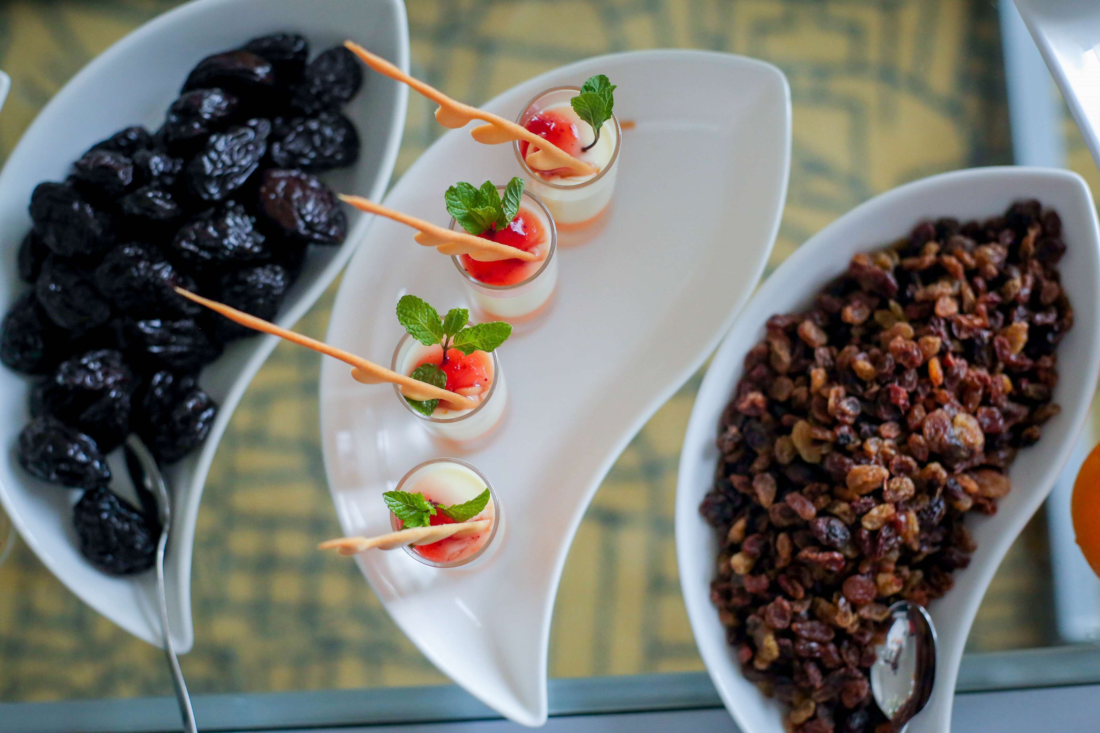 Uma mesa com três pratos: uma tigela de azeitonas pretas, um prato com cinco copinhos de sobremesa cobertos de hortelã e uma tigela de mix de frutos secos.