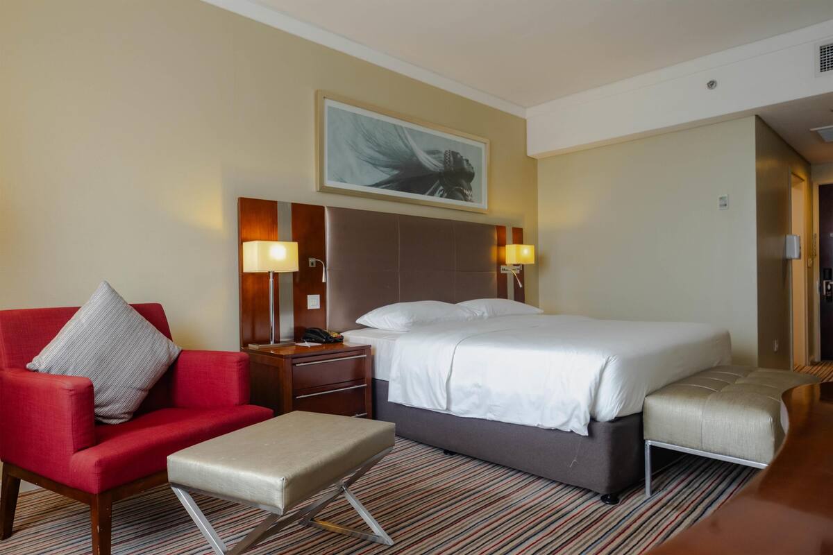 A hotel room with a bed, a red armchair, a small table, a bench, and a painting on the wall.
