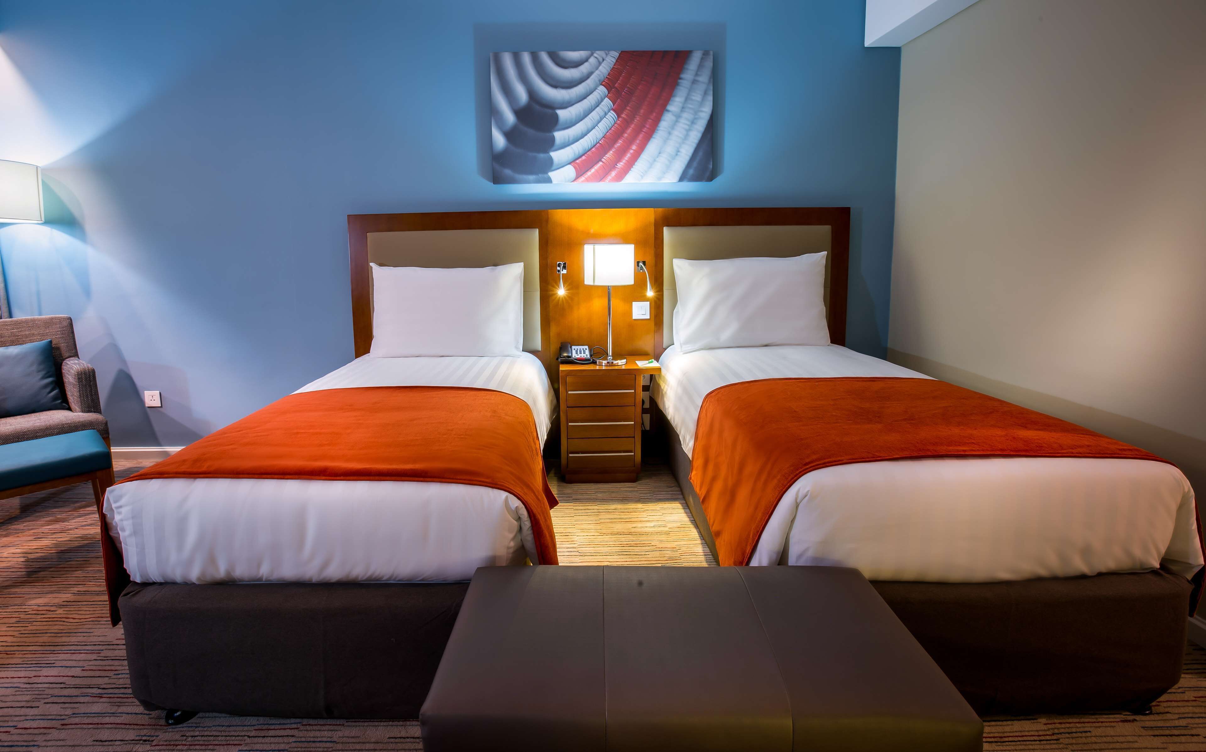 Ein Hotelzimmer mit zwei Betten, einem Nachttisch mit Lampe, einem Sessel und einem großen Bild an der Wand.