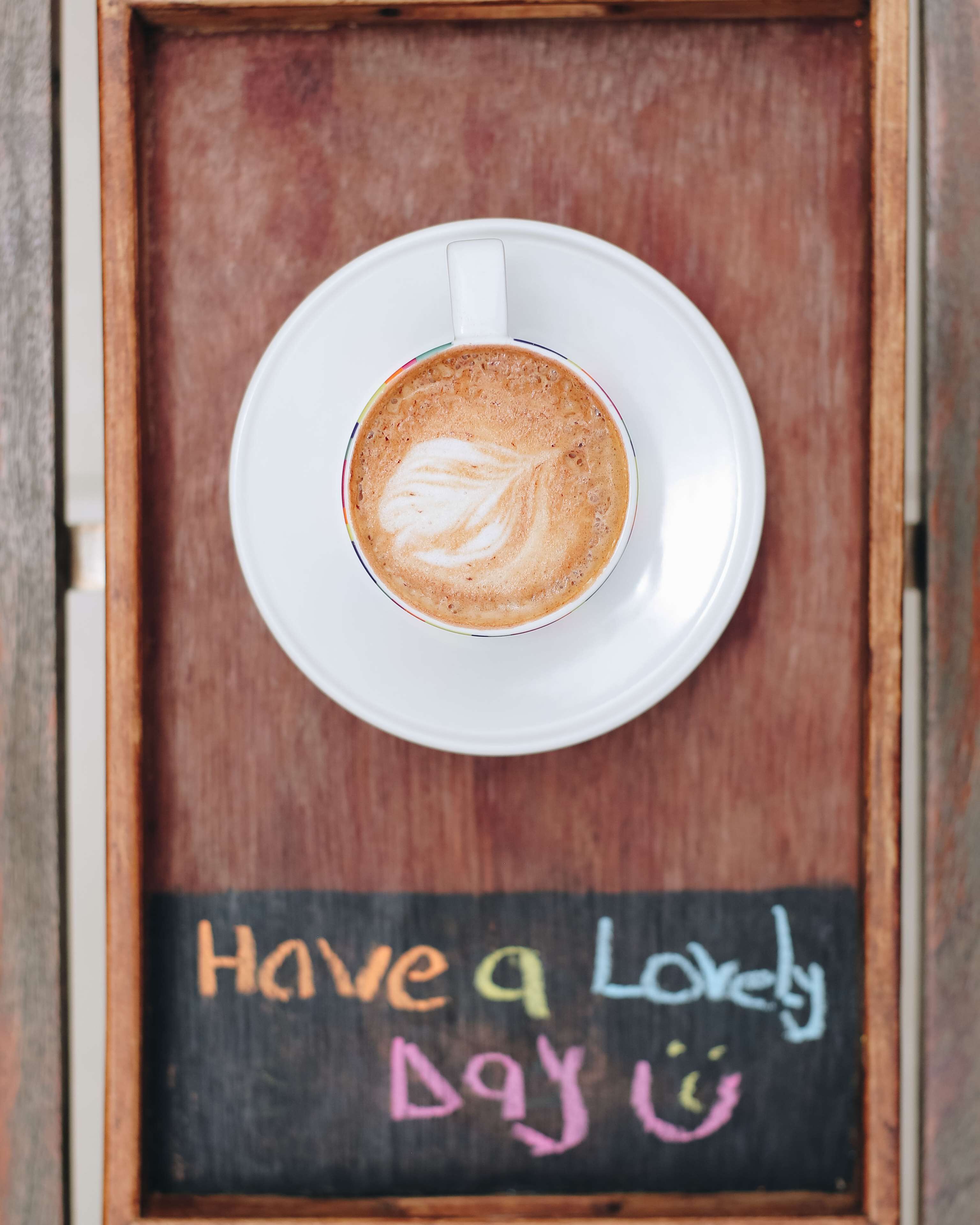 A cup of coffee with latte art on a white saucer, placed on a wooden tray with a chalkboard below it that reads "Have a Lovely Day".