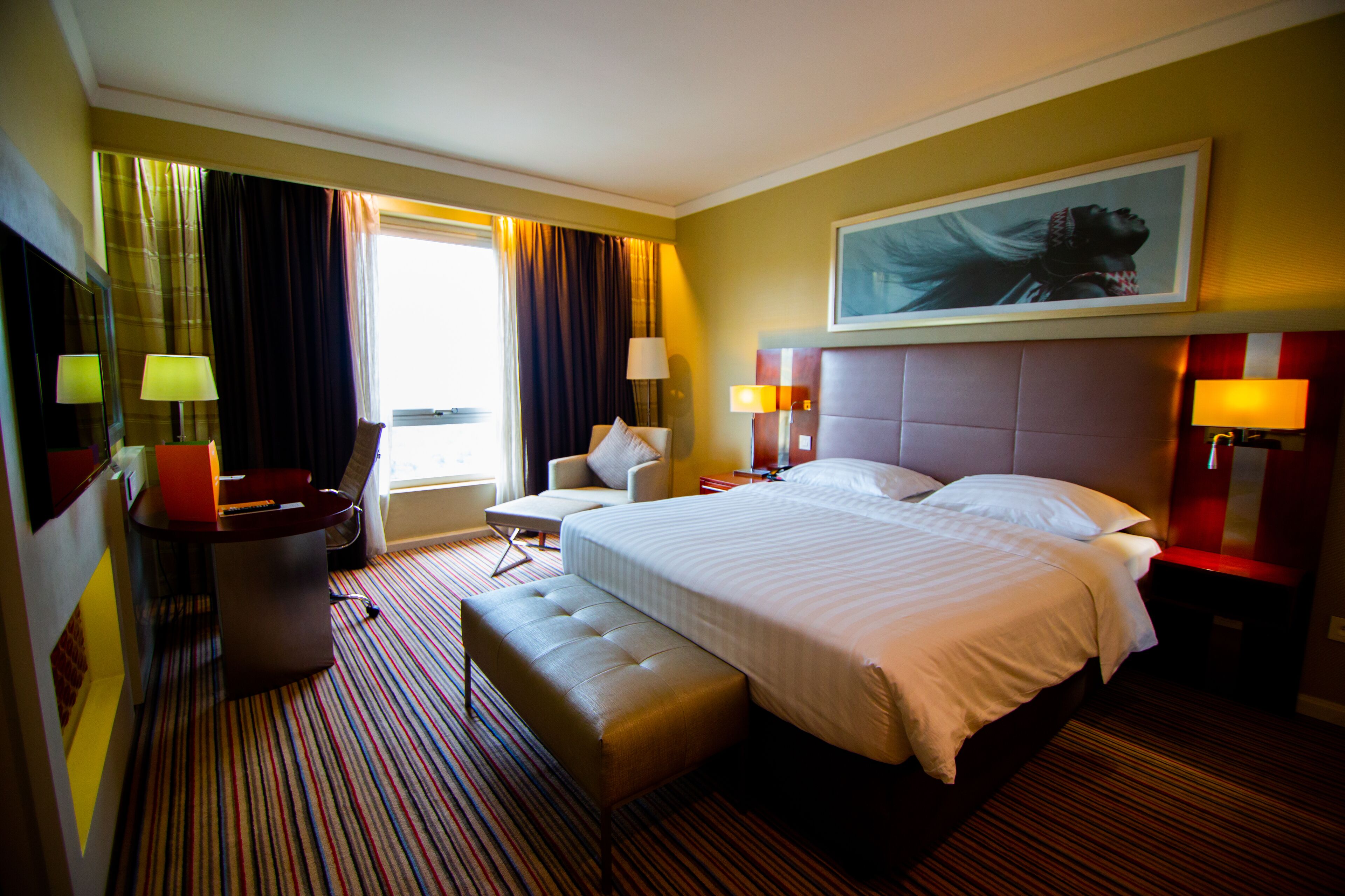 Habitación de hotel con una cama grande, una zona de estar con una silla y una mesita, un escritorio con lámpara, y un amplio ventanal con cortinas.